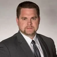 A man with short dark hair, wearing a dark suit, white shirt, and dark tie, posing against a plain light gray background.