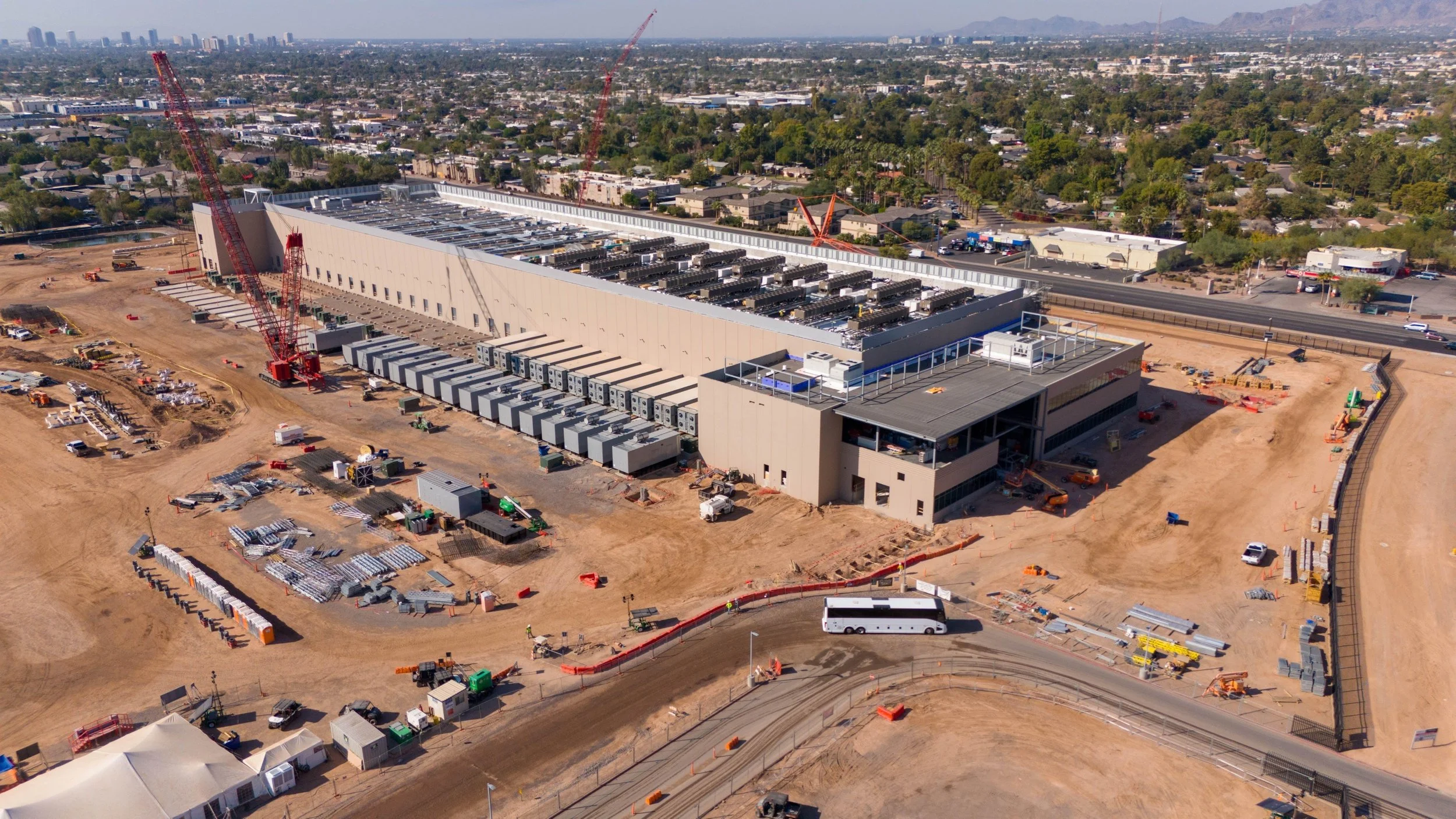 Data Center Construction. An aerial view of a large commercial building under construction, with cranes, construction equipment, and materials visible around the site, overlooking a city with mountains in the distance.