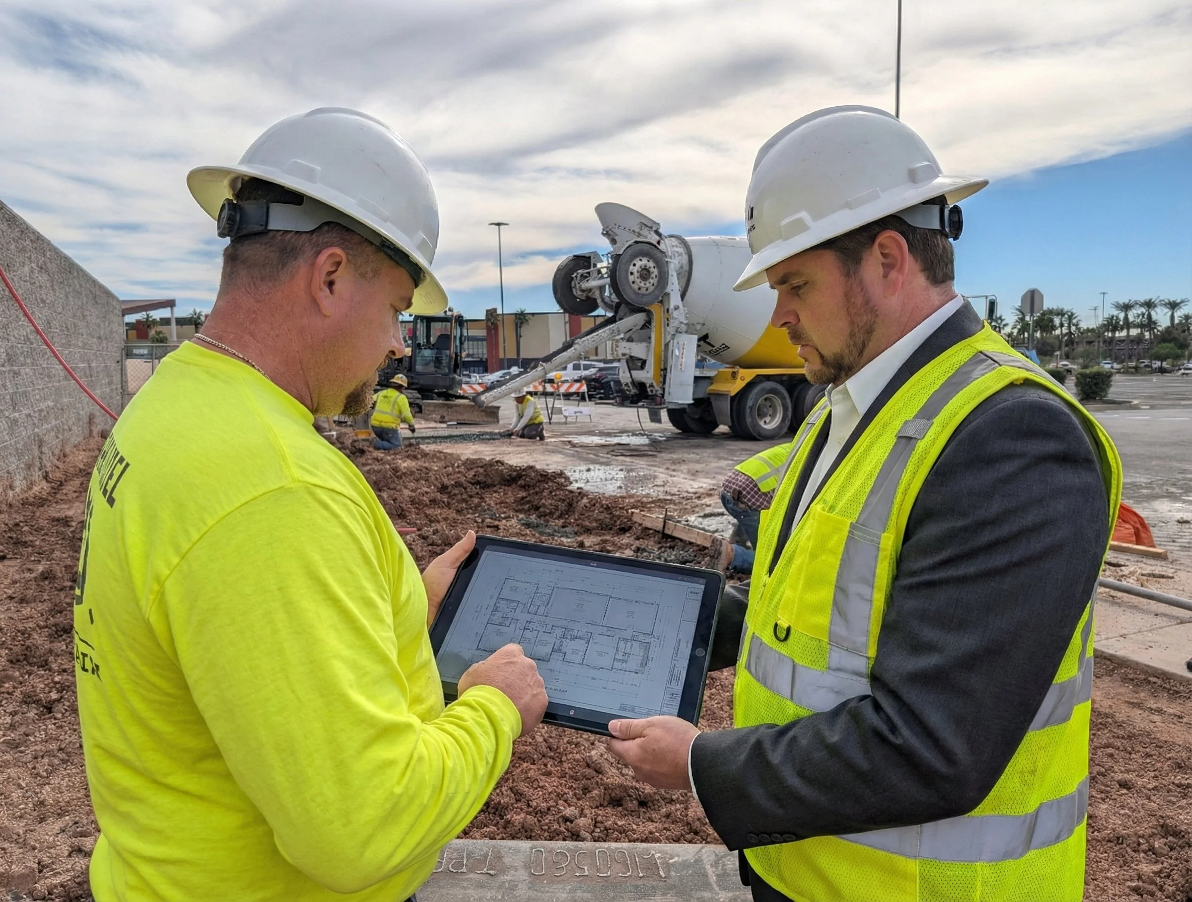 Two construction workers wearing safety helmets and high-visibility vests look at a tablet on a construction site with a concrete mixer truck and other workers in the background.
