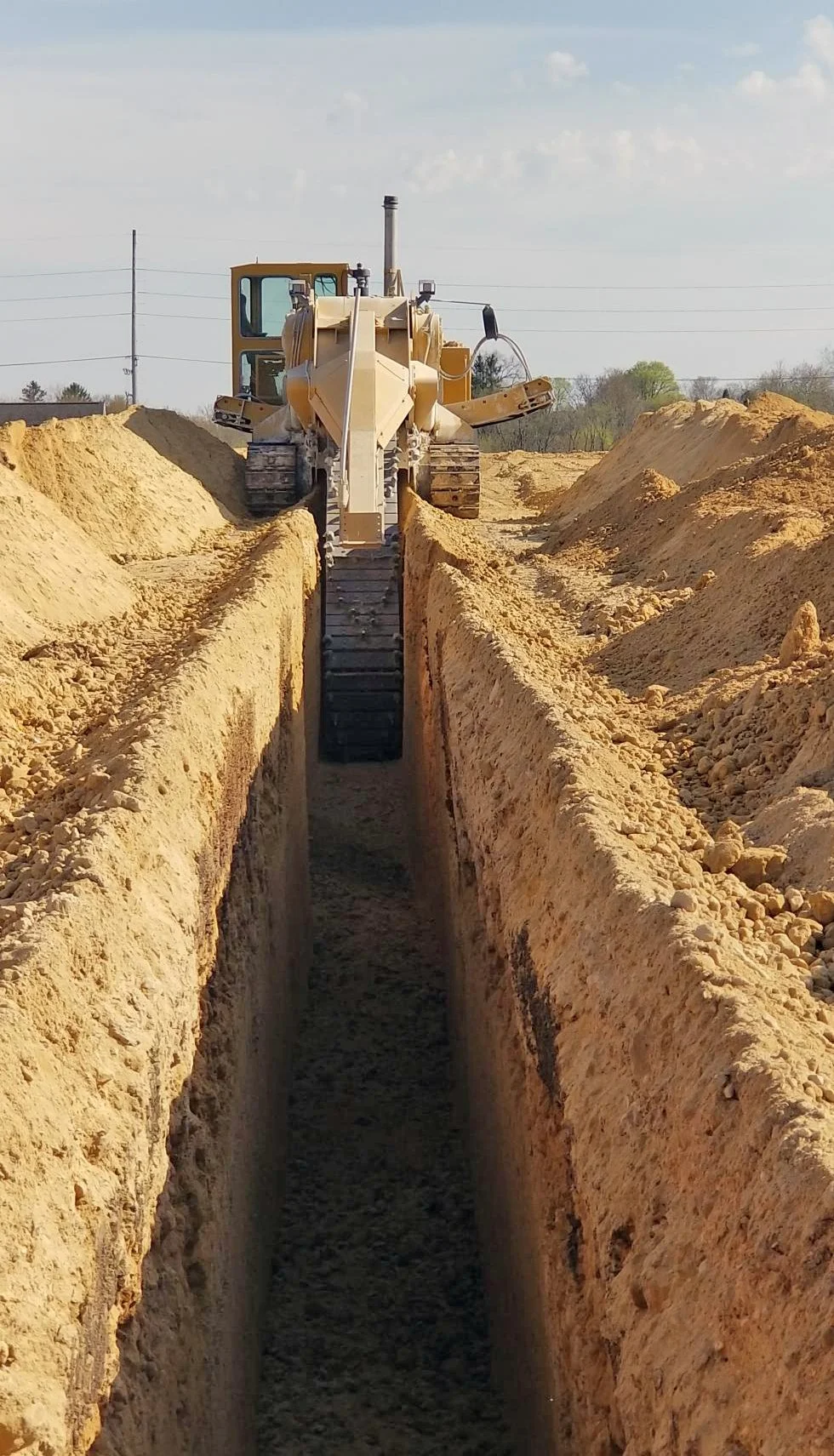 A yellow bulldozer digging a trench in a construction site with mounded dirt on either side, under a partly cloudy sky.