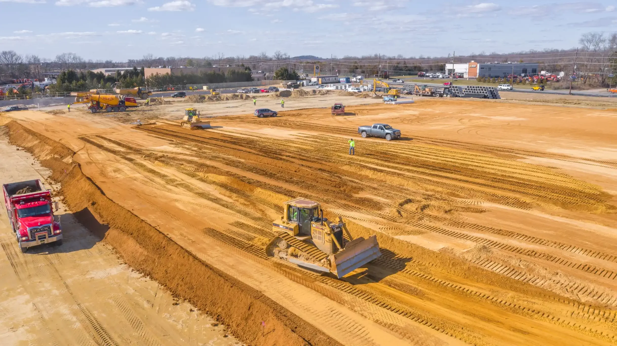 Earthwork Construction in phoenix, az with bulldozer smoothing dirt, trucks, and scattered construction workers, with a nearby highway and buildings in the background.