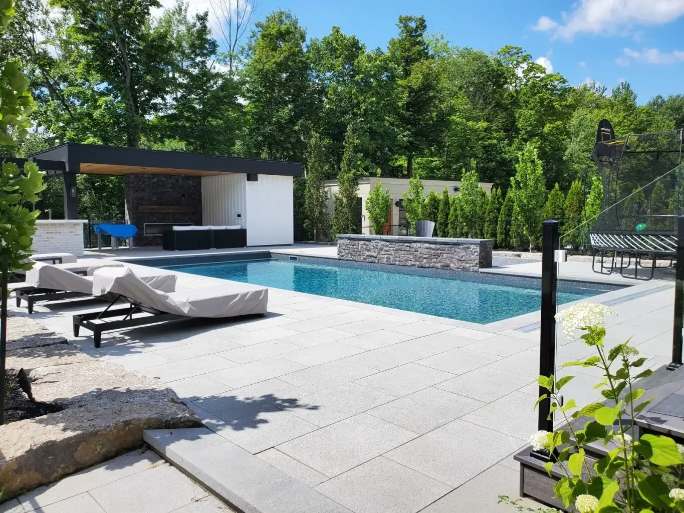 Modern backyard with a swimming pool, two lounge chairs, a shaded patio area, and lush green trees in the background.
