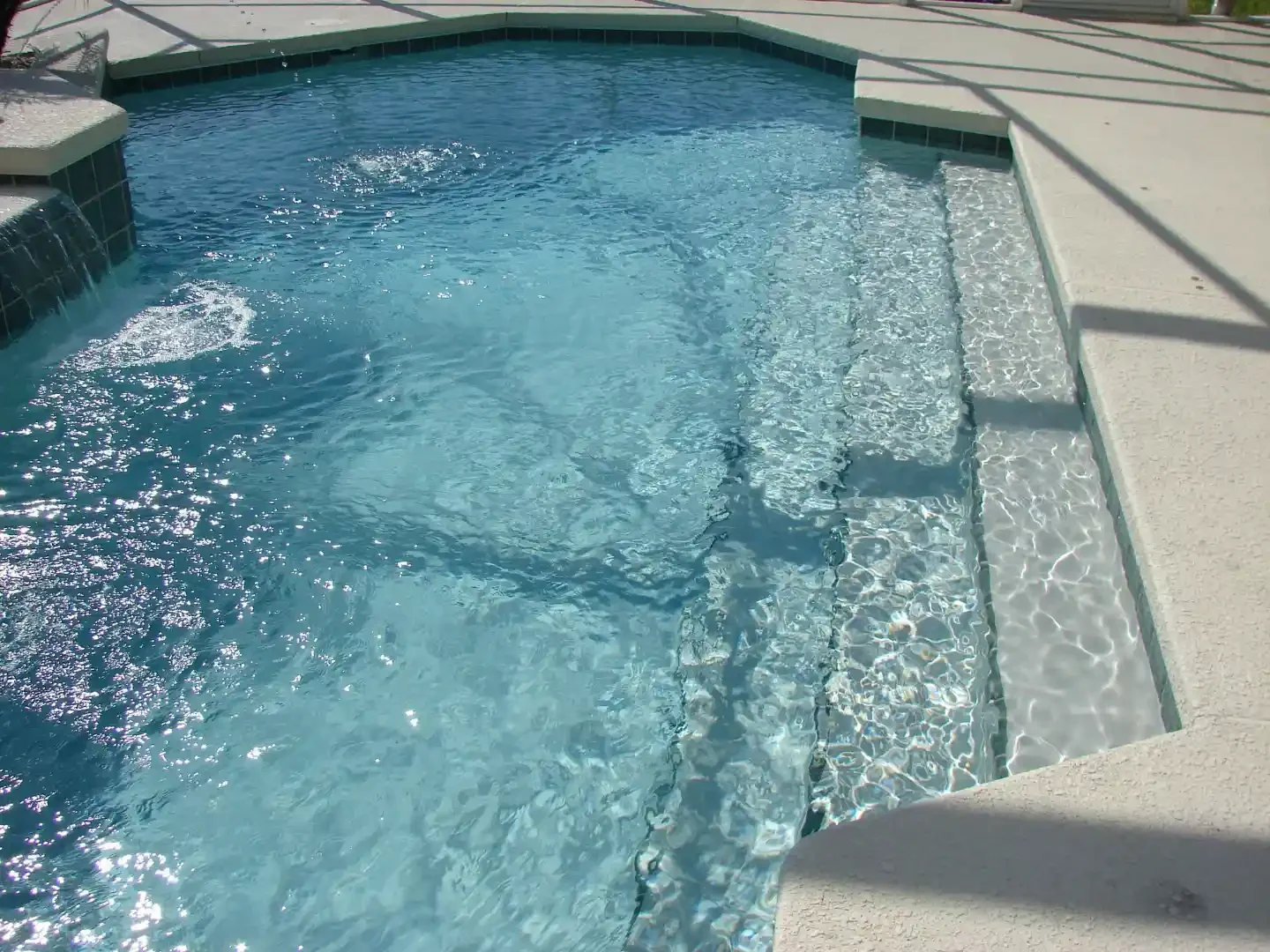 A clear blue swimming pool with steps leading into the water, surrounded by a concrete deck and fencing.