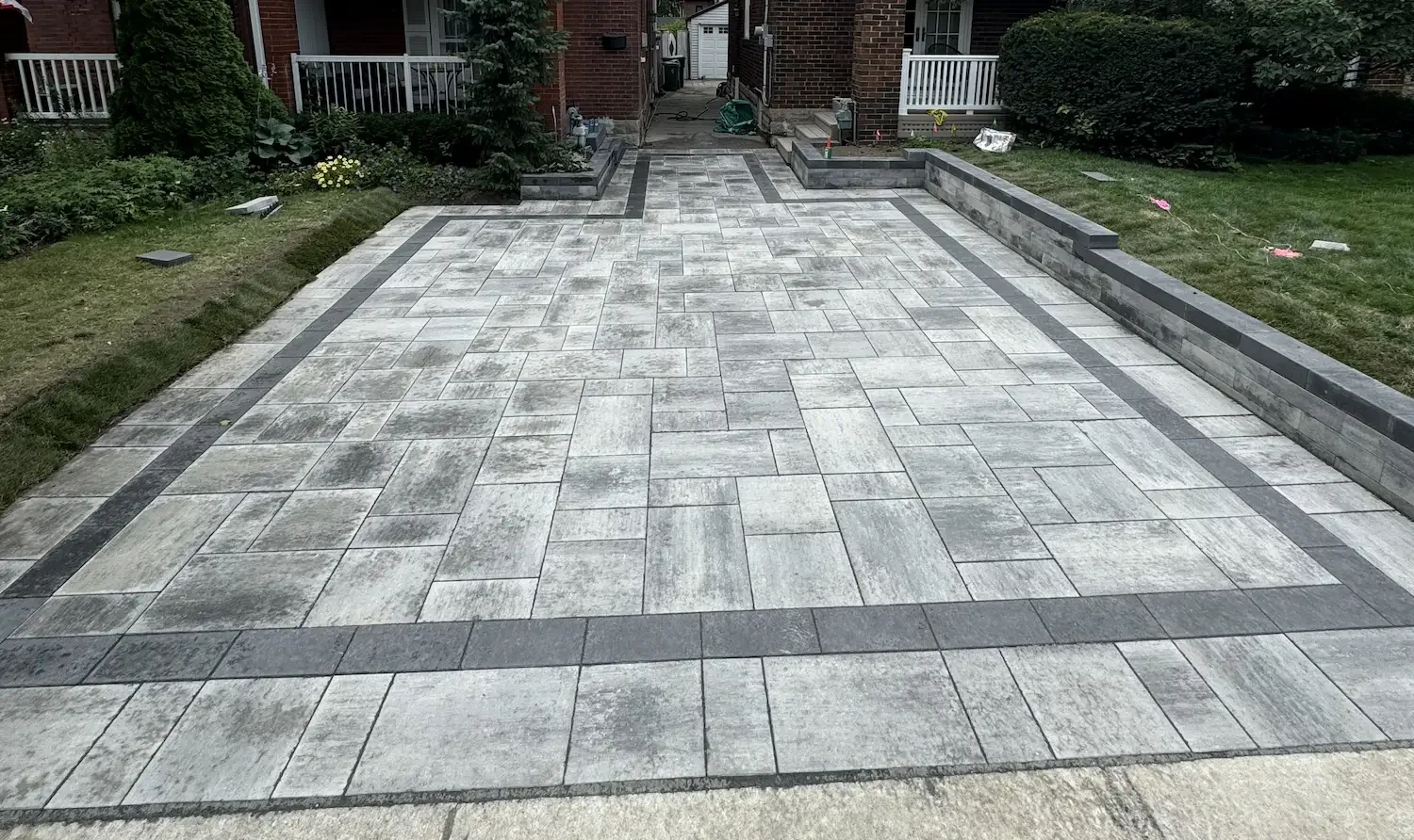 A newly paved driveway with light and dark gray bricks arranged in a pattern, bordered by darker bricks, leading to brick houses with front porches and surrounded by green grass and shrubs.