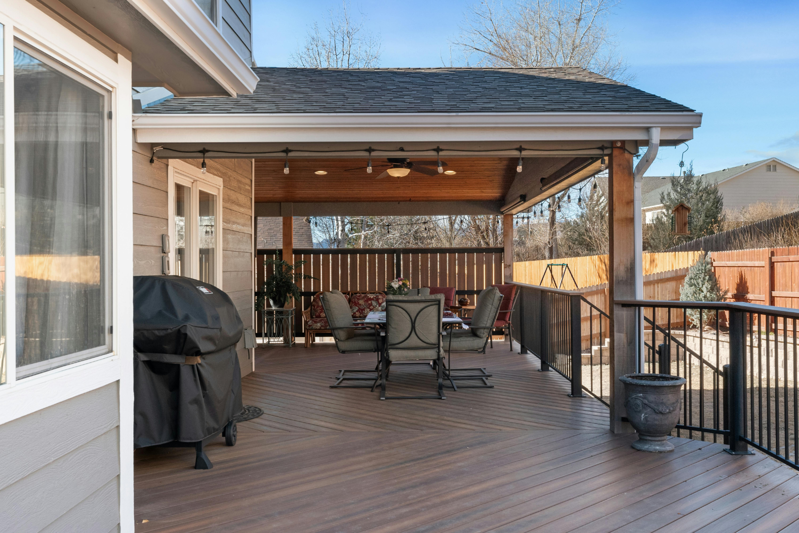 A spacious backyard deck with outdoor furniture, a grill, string lights, and a fenced yard under a clear blue sky.