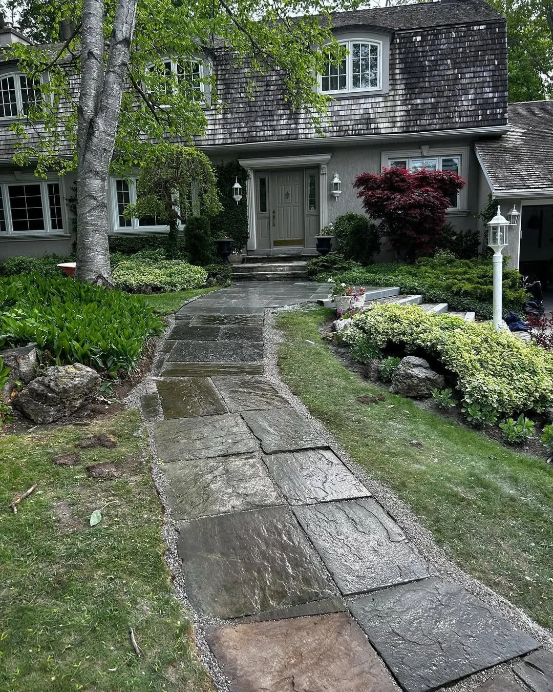 A stone pathway leading to a house entrance surrounded by lush green plants, trees, and garden decor.