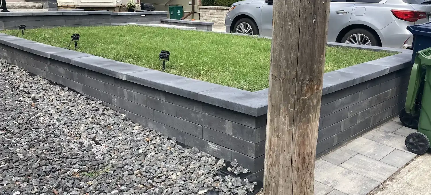 Image of a raised garden bed made of gray bricks with green grass inside, surrounded by gravel and stone pavers, with parked cars and a wooden utility pole nearby.