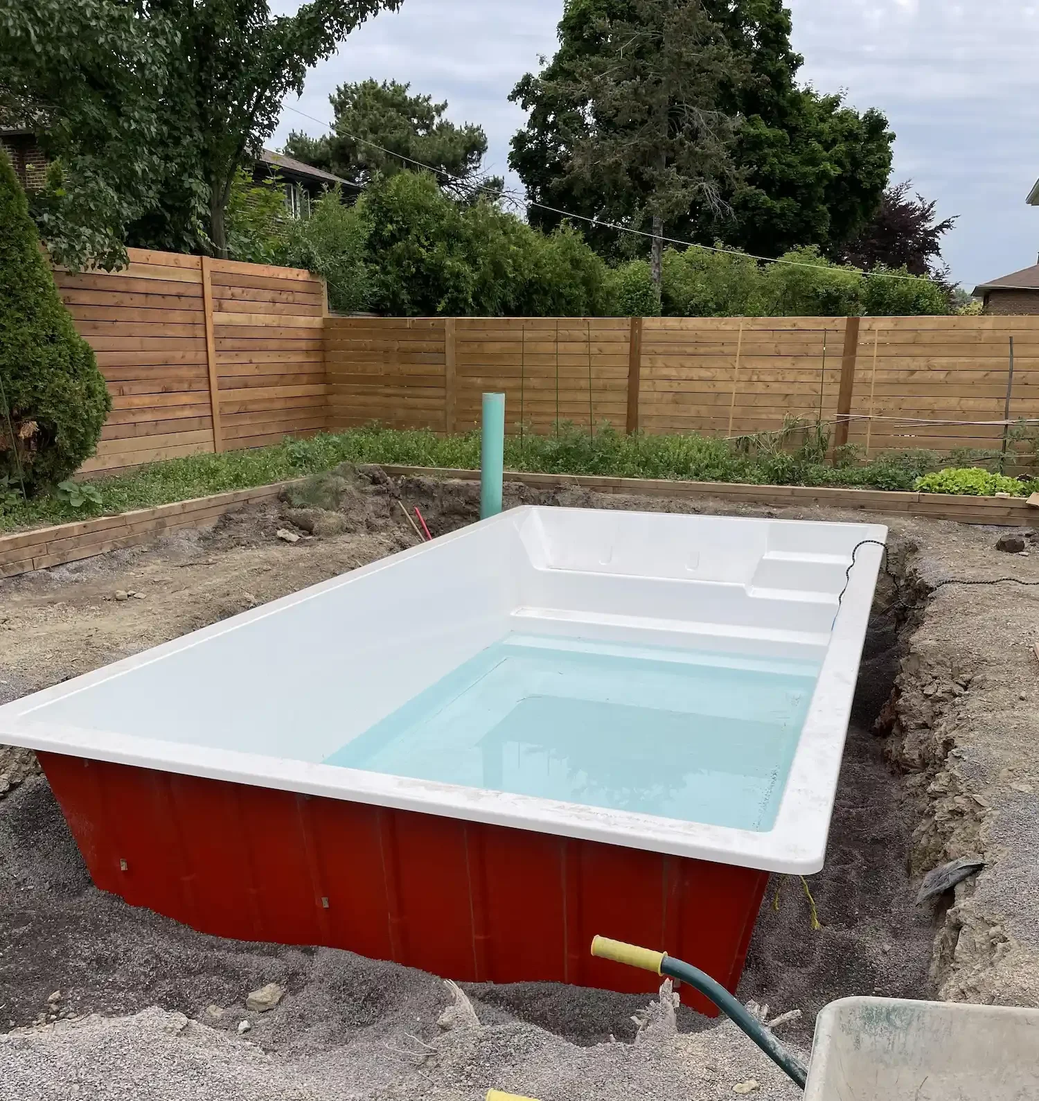 A backyard area under construction with a partially installed rectangular hot tub filled with water, surrounded by dirt and construction materials, enclosed by a wooden privacy fence, with trees and neighboring houses in the background.