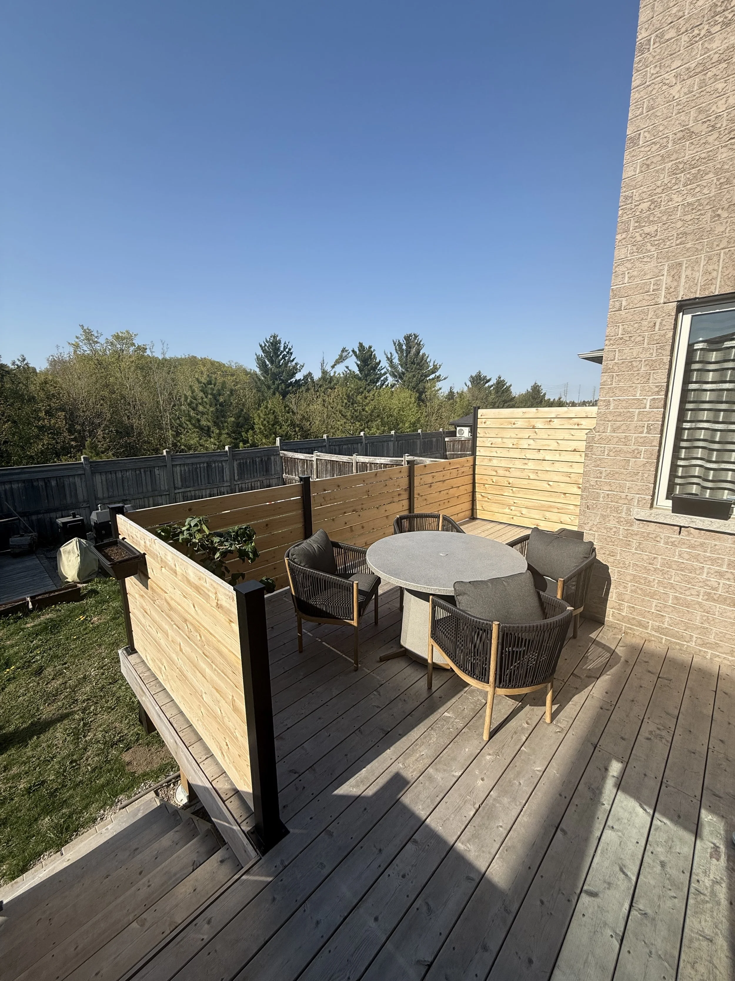 Empty outdoor wooden deck with a round table and four chairs, surrounded by a wooden fence, with trees in the background and a brick house wall on the right side.
