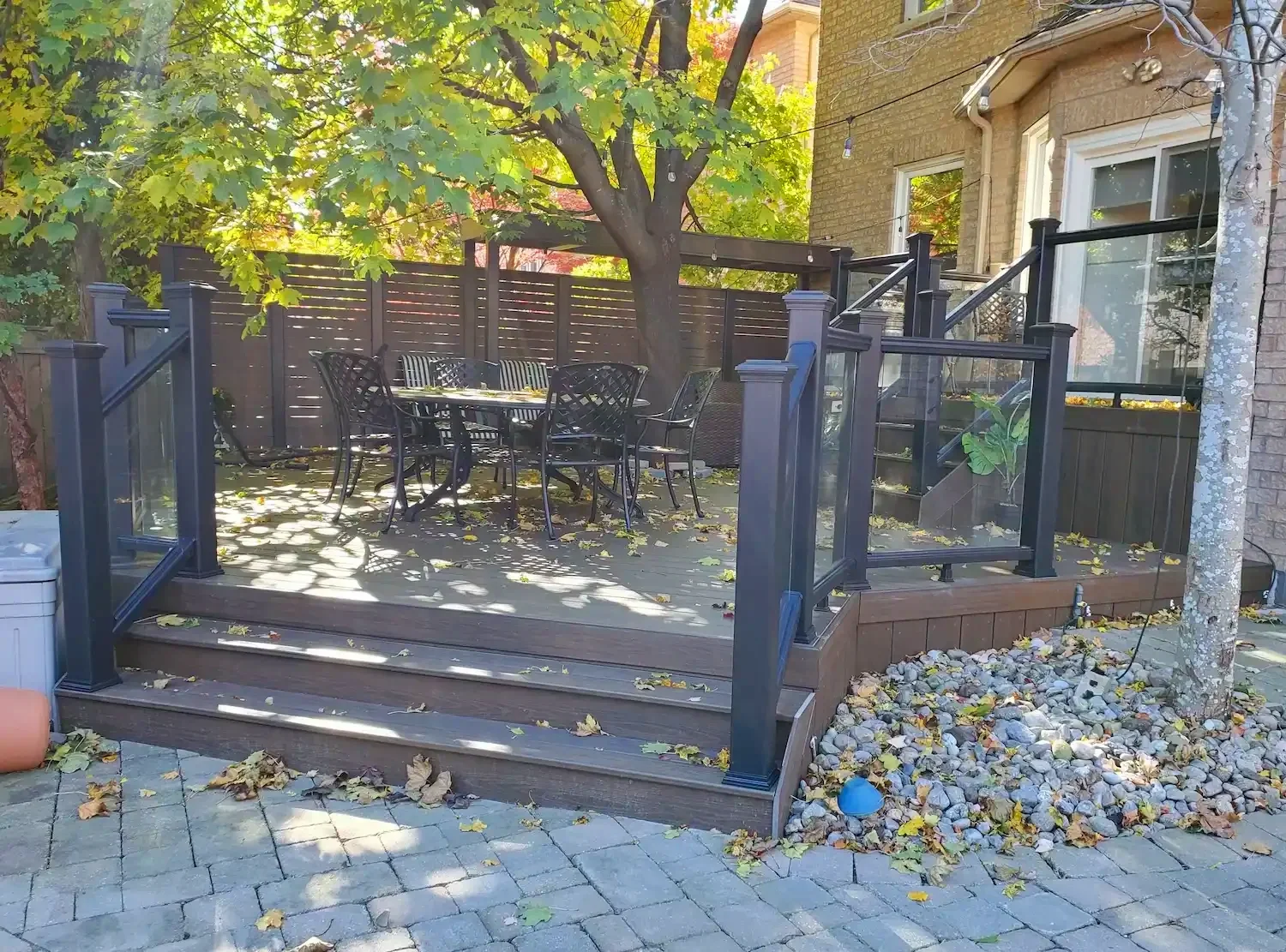 Backyard deck with black railing, outdoor table and chairs, surrounded by trees with green leaves, fallen leaves on the deck and ground, and a brick house in the background.