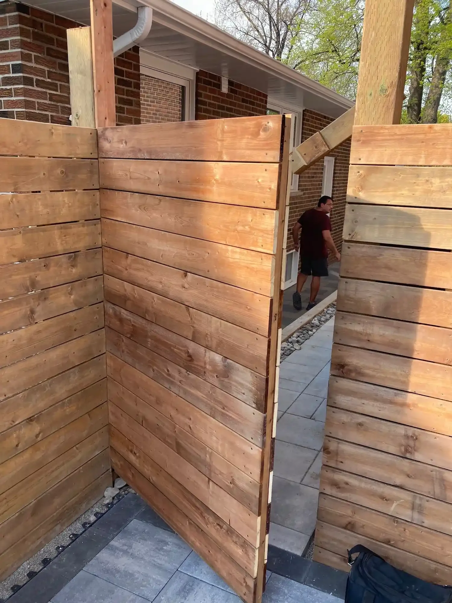 A person walking inside a small outdoor area with wooden fencing, brick house walls, and a tiled pathway.