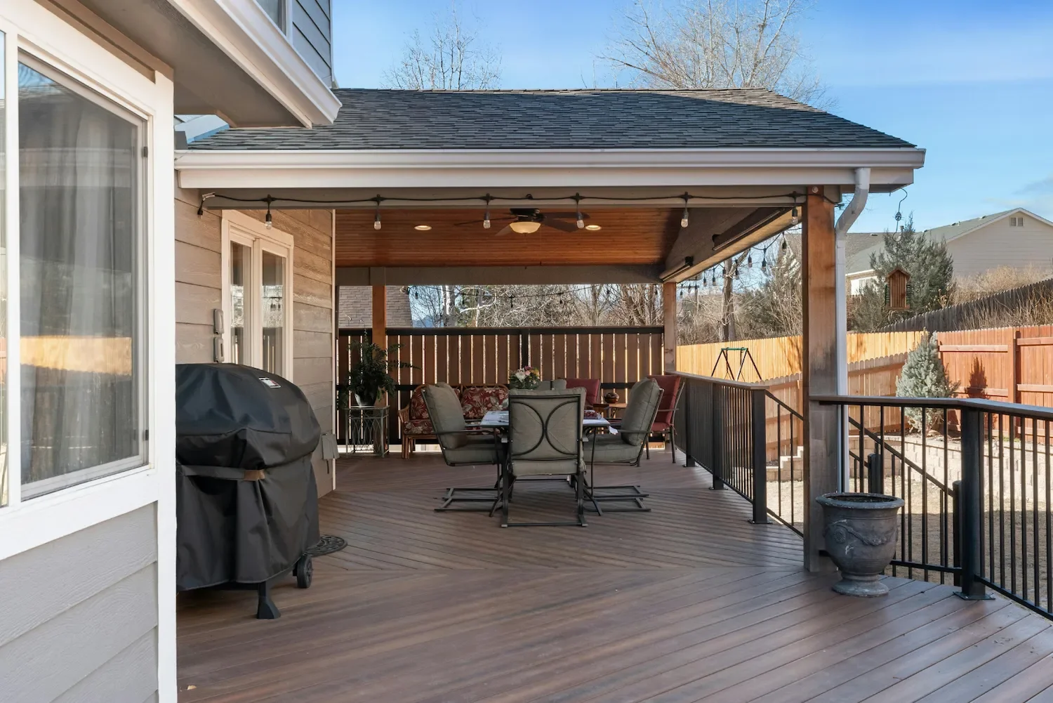 Empty backyard patio with wooden deck, covered ceiling, outdoor dining table with chairs, potted plants, a barbecue grill, and fencing with a swing set in the background.