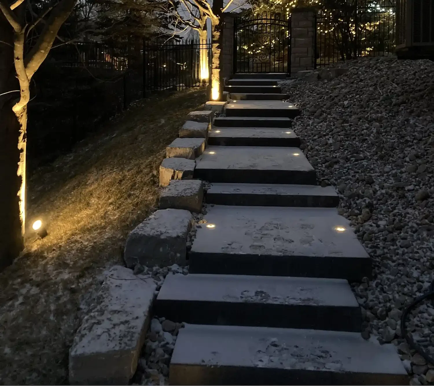 Snow-covered stone stairs leading up to a gate, illuminated by ground lights at night.