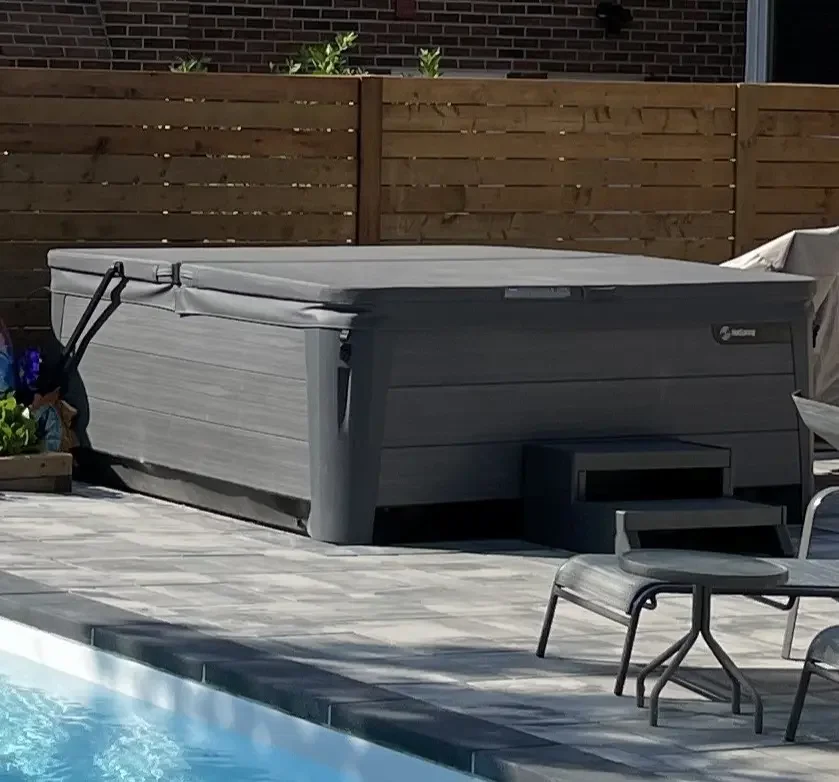 A backyard with a closed hot tub cover next to a swimming pool, surrounded by a wooden fence and patio furniture.