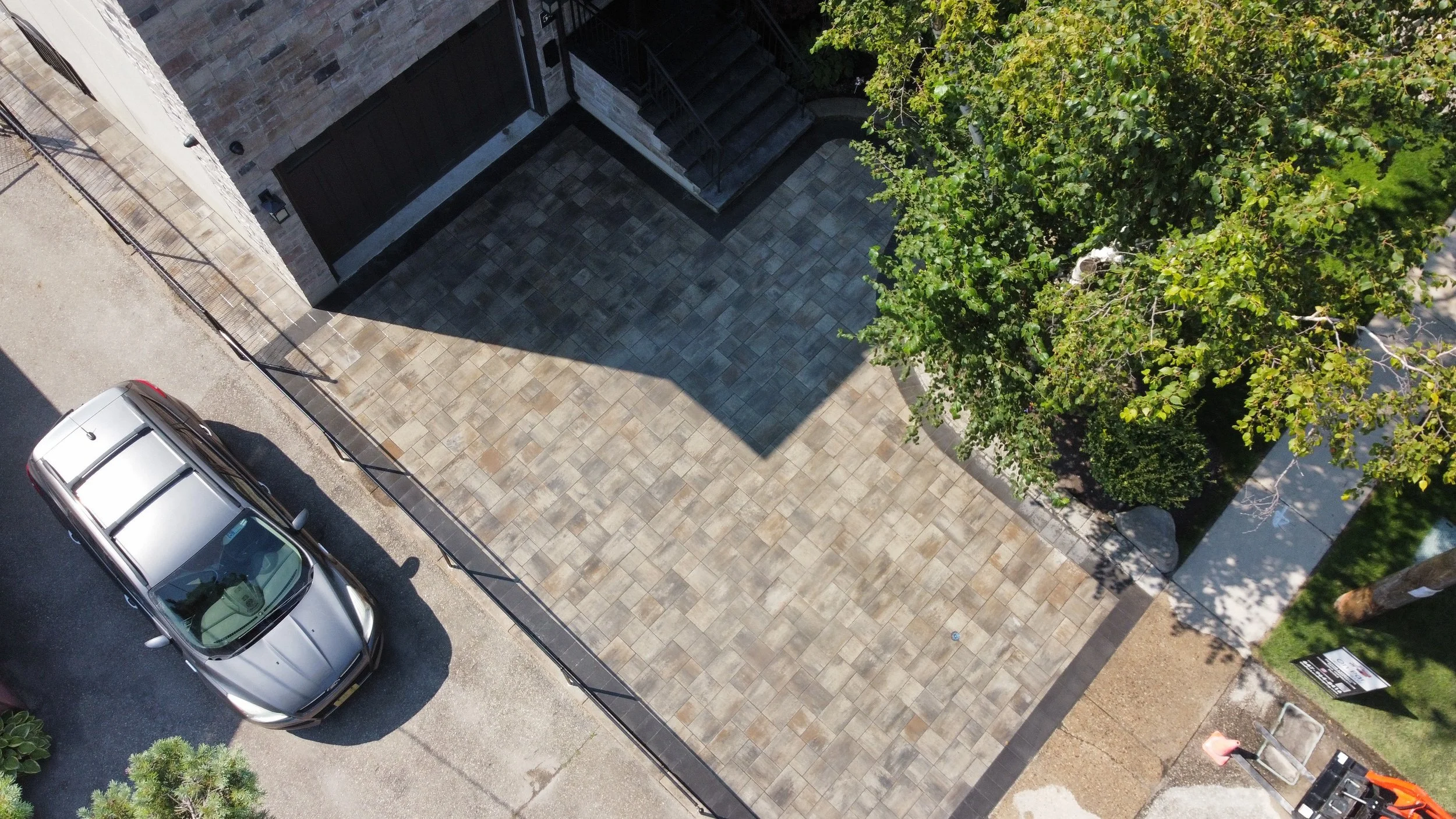 Aerial view of a driveway with brick paving, a parked silver car, an adjacent building with stairs, and a tree with green foliage.