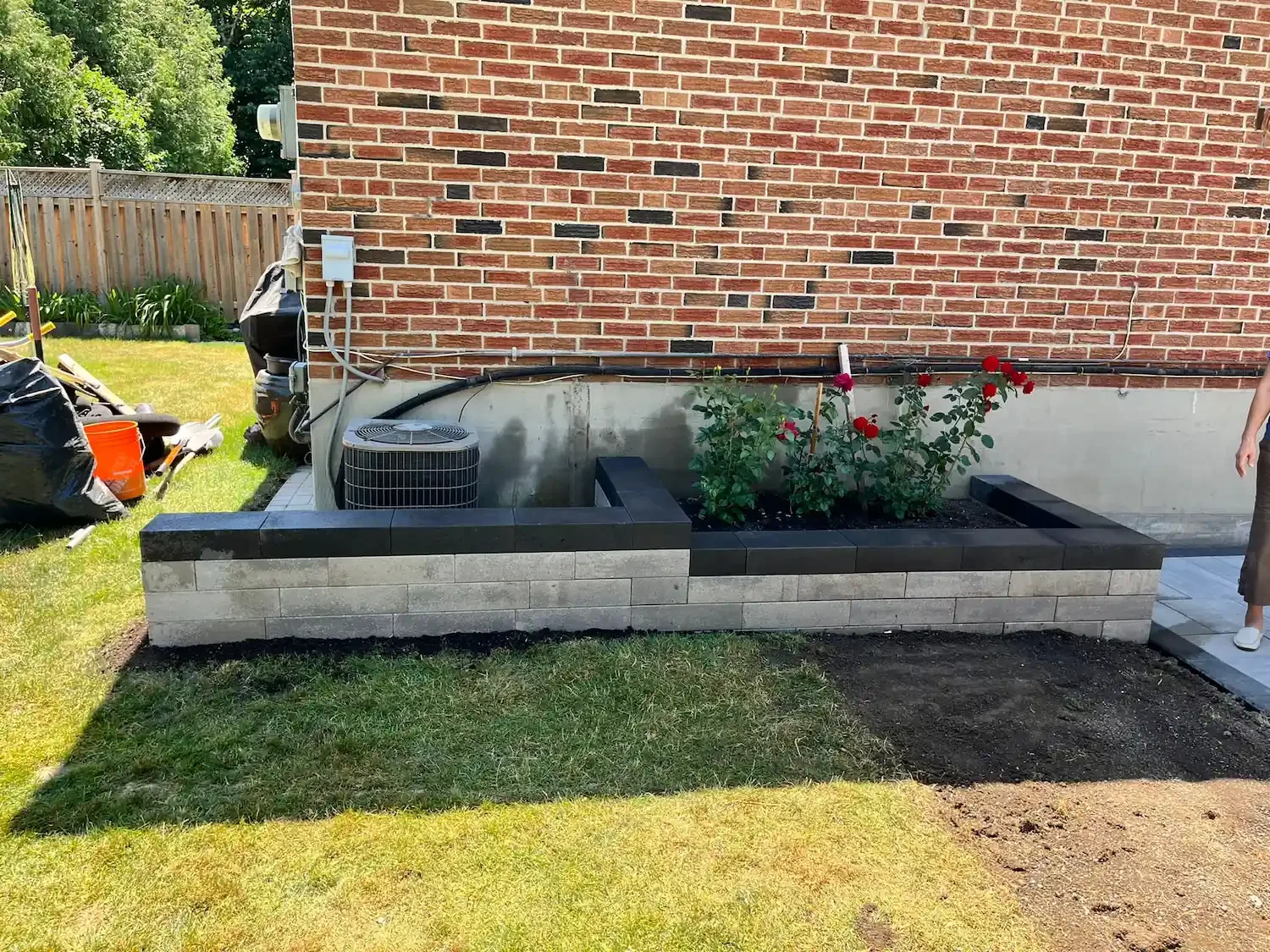 A brick house exterior with a garden bed containing red roses, a paved walkway, and a lawn. Gardening tools and equipment are on the left, and a person's leg is visible on the right.
