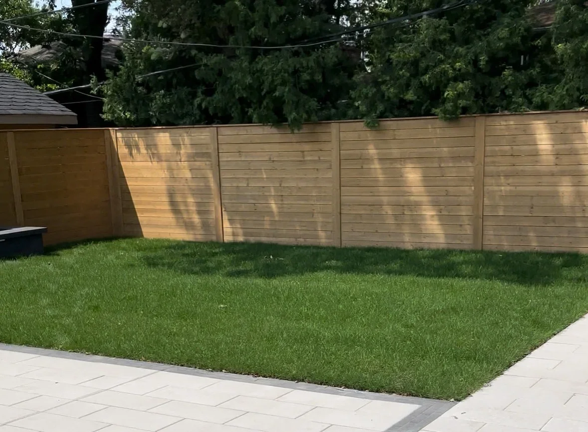 Backyard with a green lawn, a wooden privacy fence, part of a patio, and a large tree with shadows cast on the fence.
