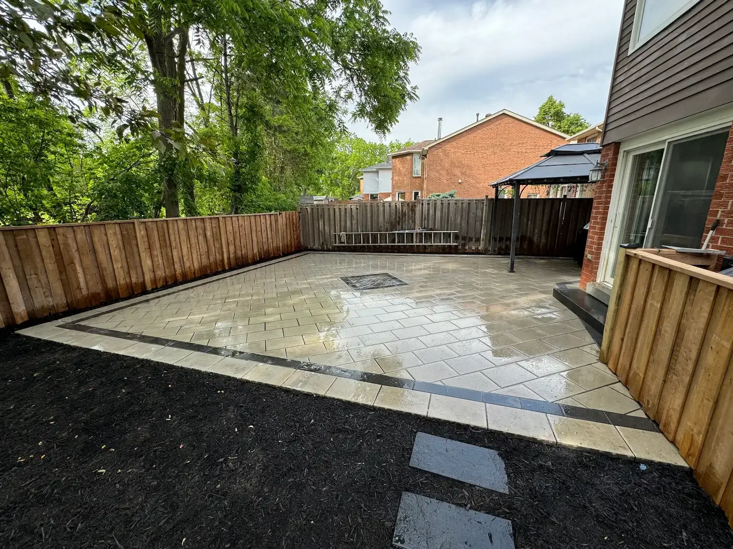 Newly paved backyard patio with wooden fencing, some trees, and neighboring houses, during a rainy day.