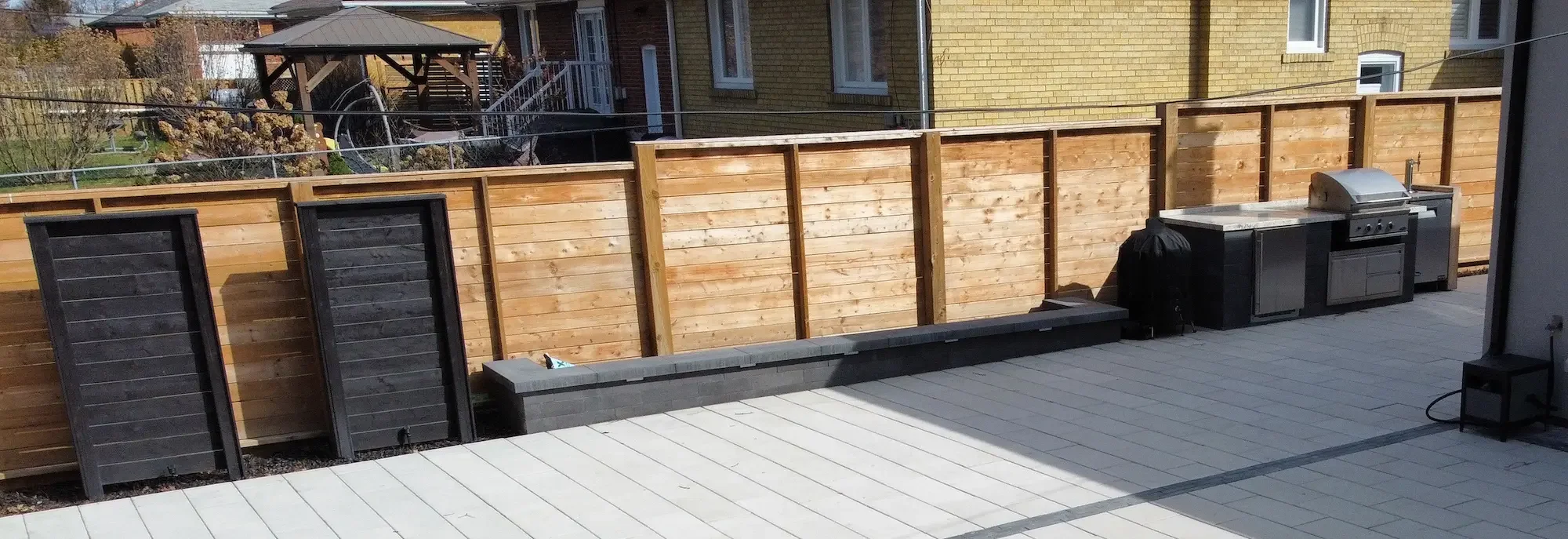 A patio with a wooden privacy fence, outdoor grill, and potted plant in black bags. The fence has surrounding houses and a small outdoor gazebo in the background.