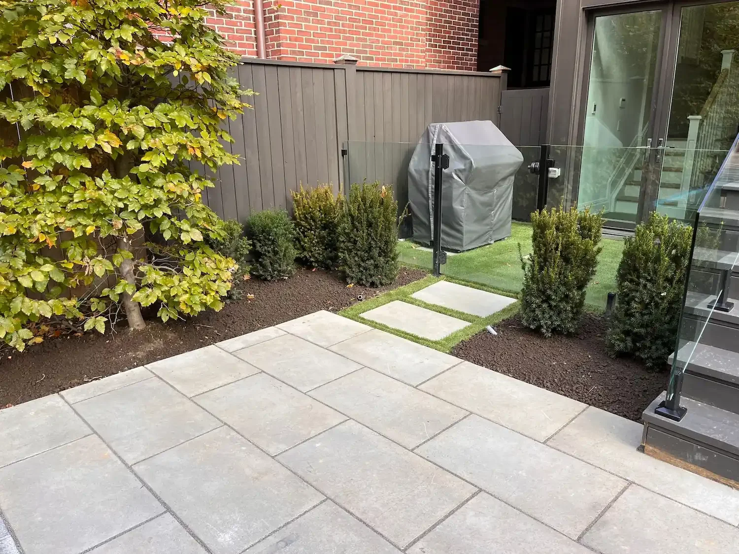 Backyard patio with large concrete tiles, small shrub plants, a gray privacy fence, a grill covered with a gray cover, glass railing, and steps leading up to a house.
