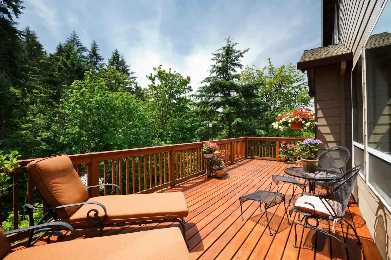A spacious wooden deck with outdoor furniture, potted plants, and hanging flower baskets, overlooking a lush green forest.