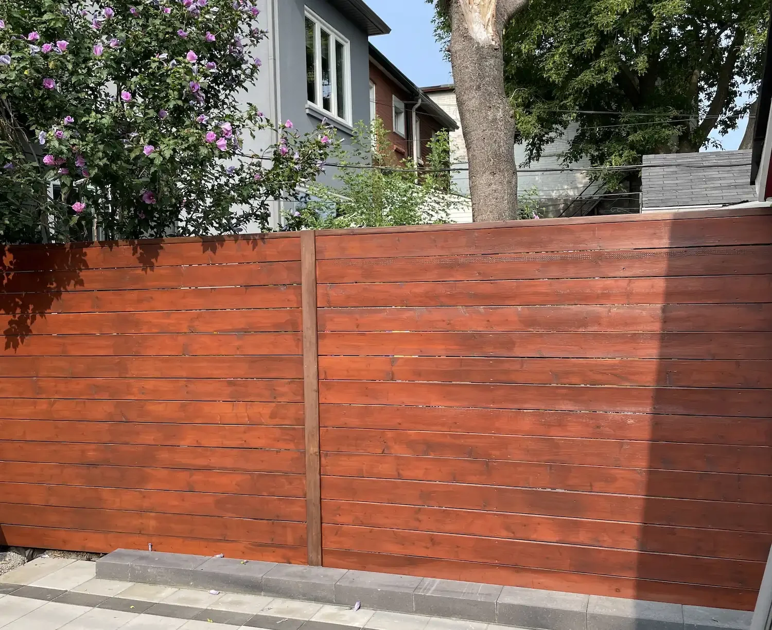 A wooden fence with horizontal planks, in front of a garden with flowering bushes and trees behind it, in a neighborhood with houses visible in the background.