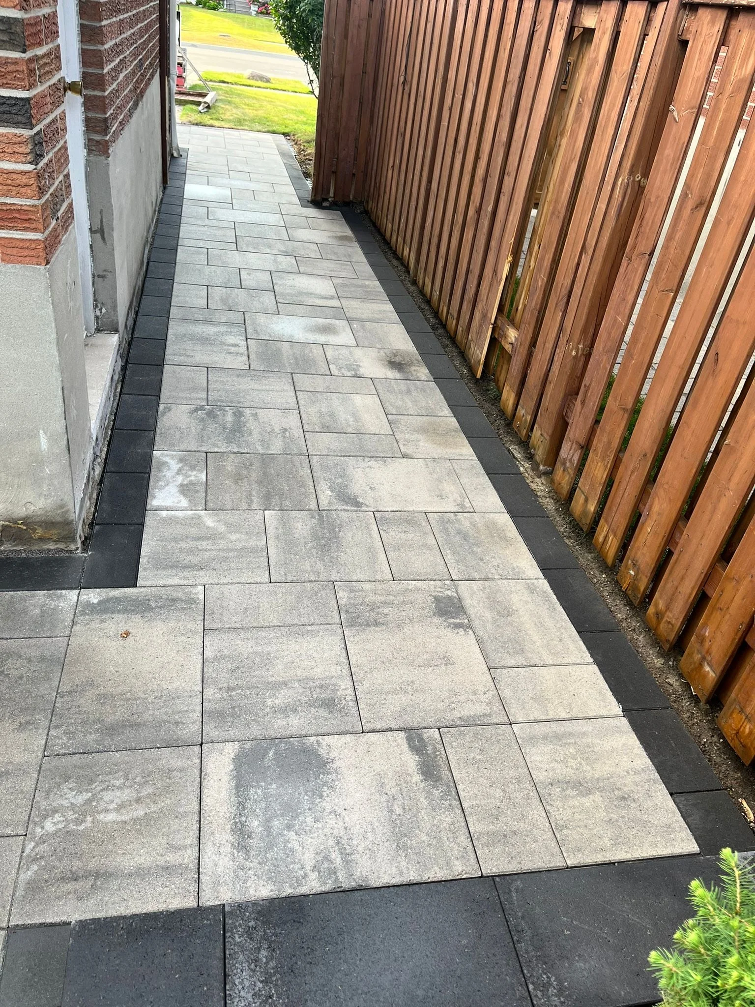 A narrow outdoor sidewalk with grey and black paving stones, bordered by a wooden fence on the right and the side of a brick building on the left, leading to a street with green grass and trees in the background.