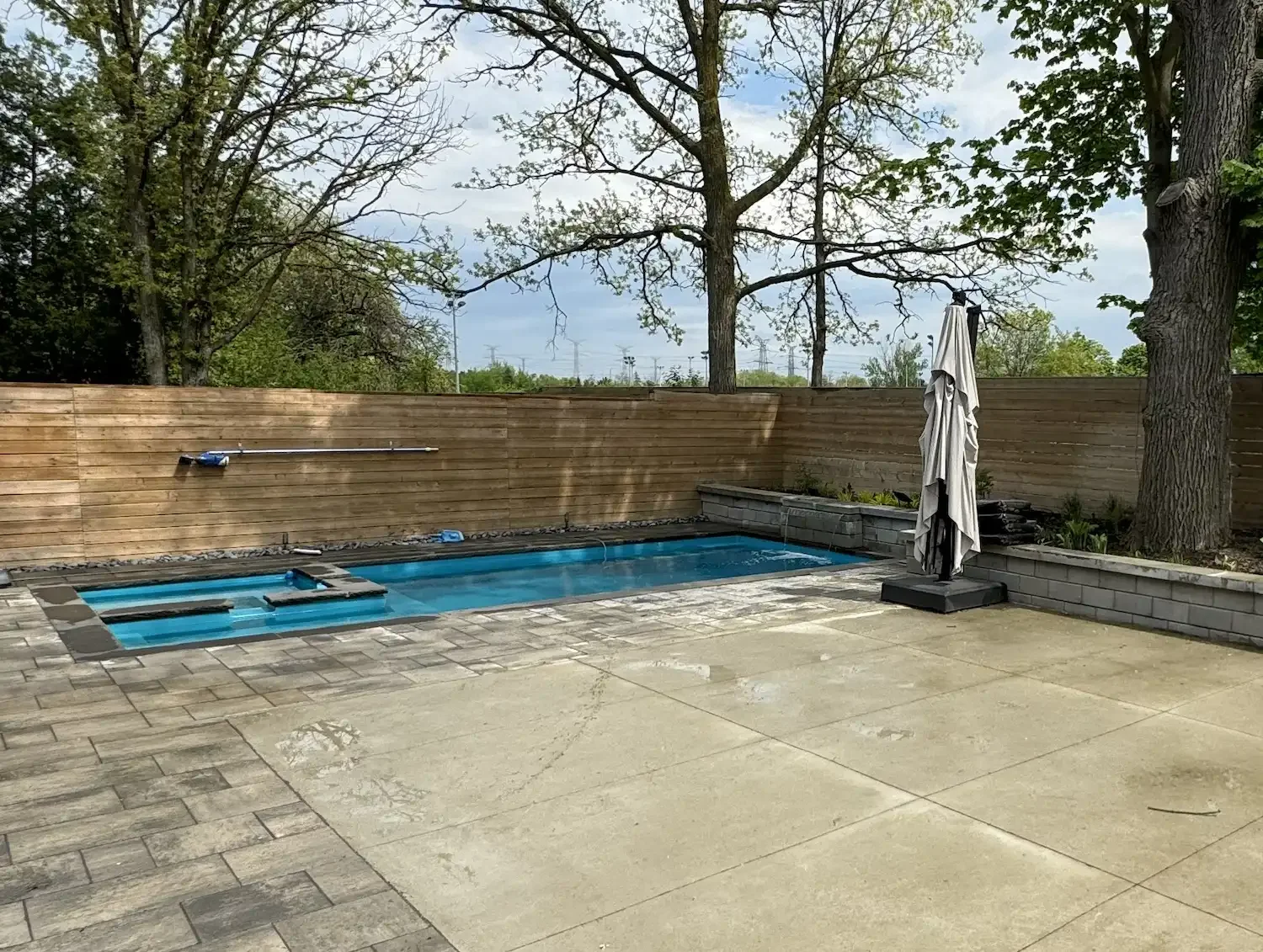 Backyard pool area with a covered hot tub, a wooden privacy fence, a closed patio umbrella, and some trees and greenery in the background.