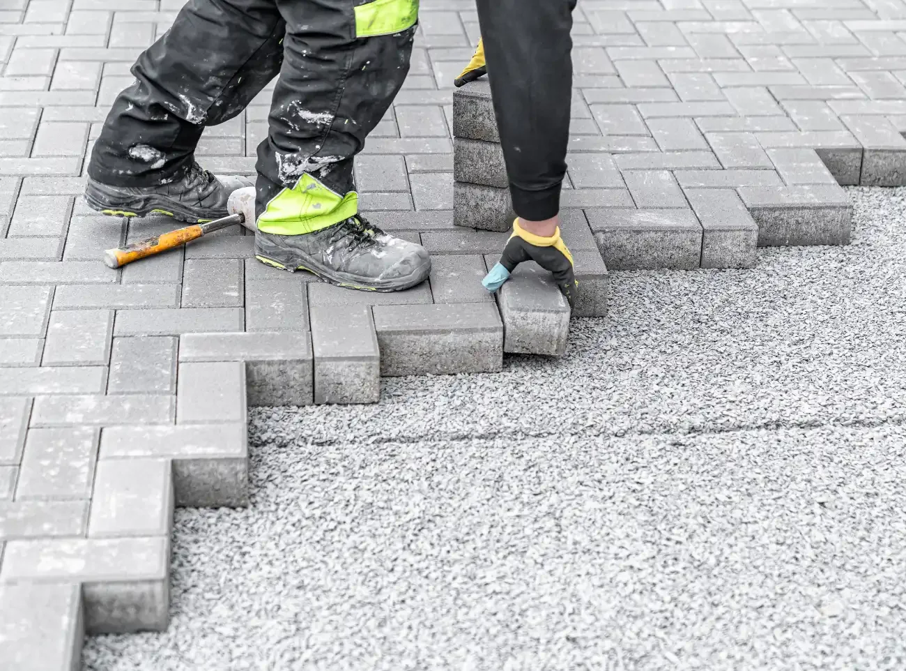 Two workers laying gray paving stones over a gravel base to create a paved surface.