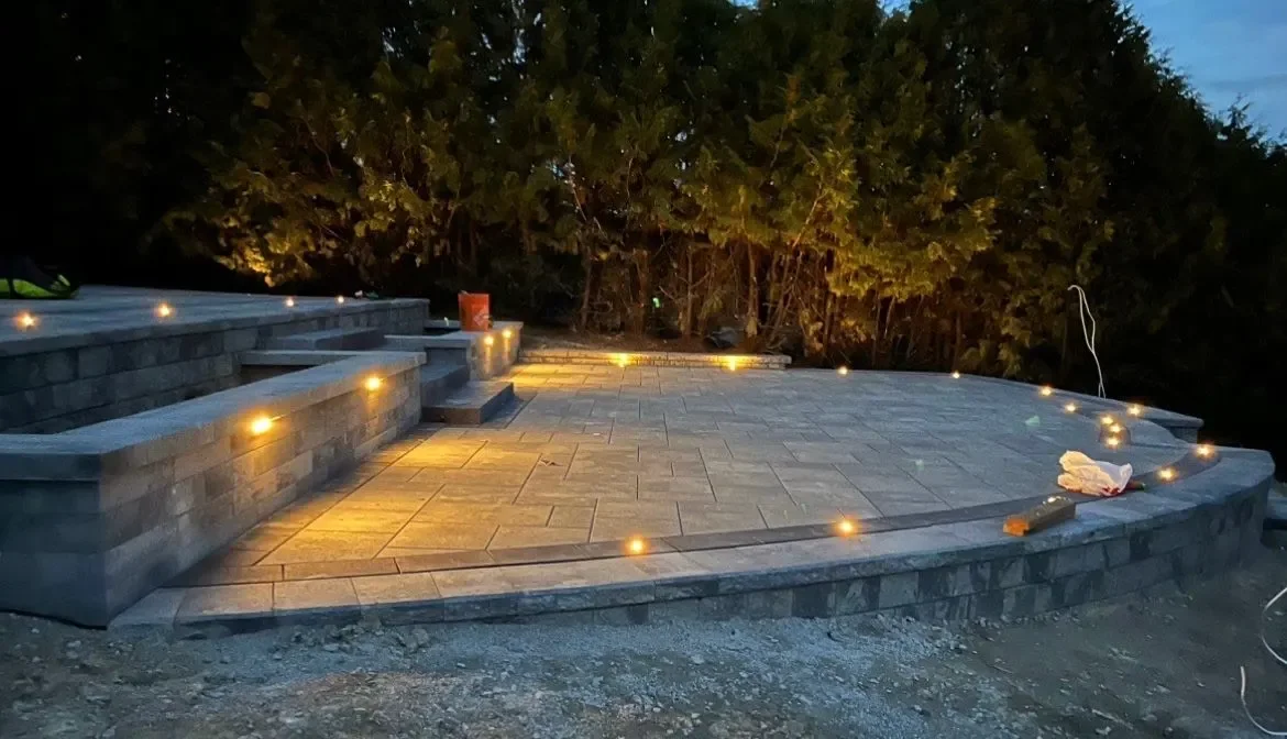 An illuminated outdoor stone patio with stairs, surrounded by trees at dusk.