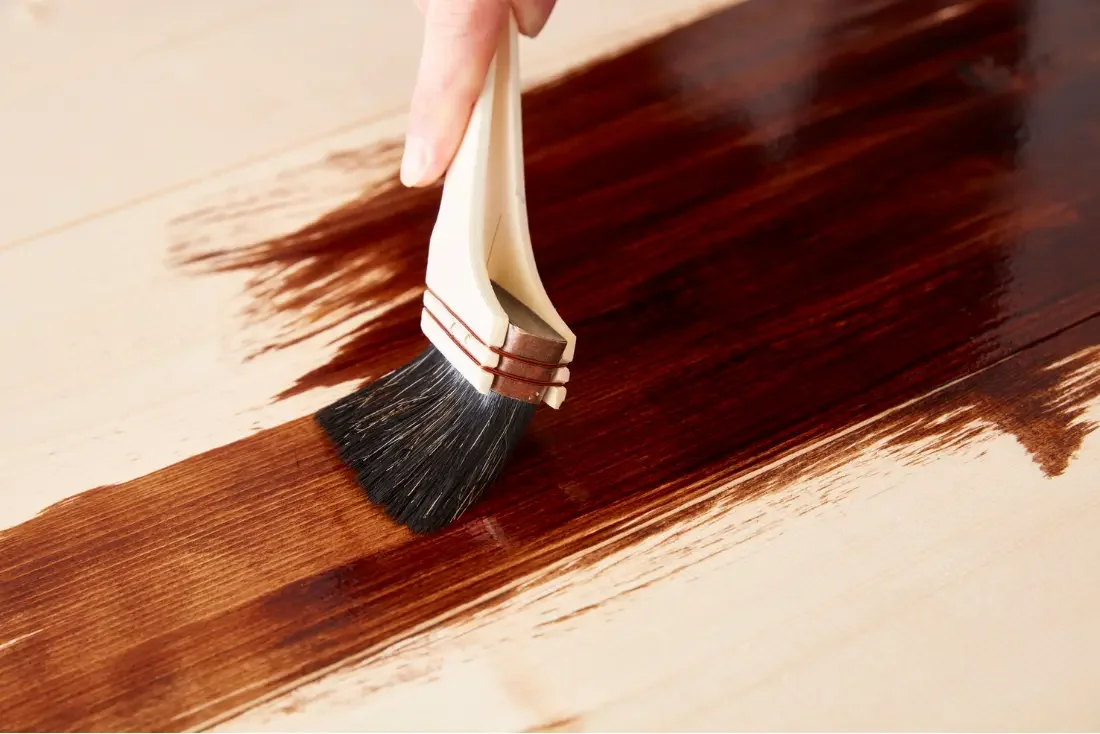 Close-up of a hand applying dark brown stain to a wooden surface with a paintbrush.