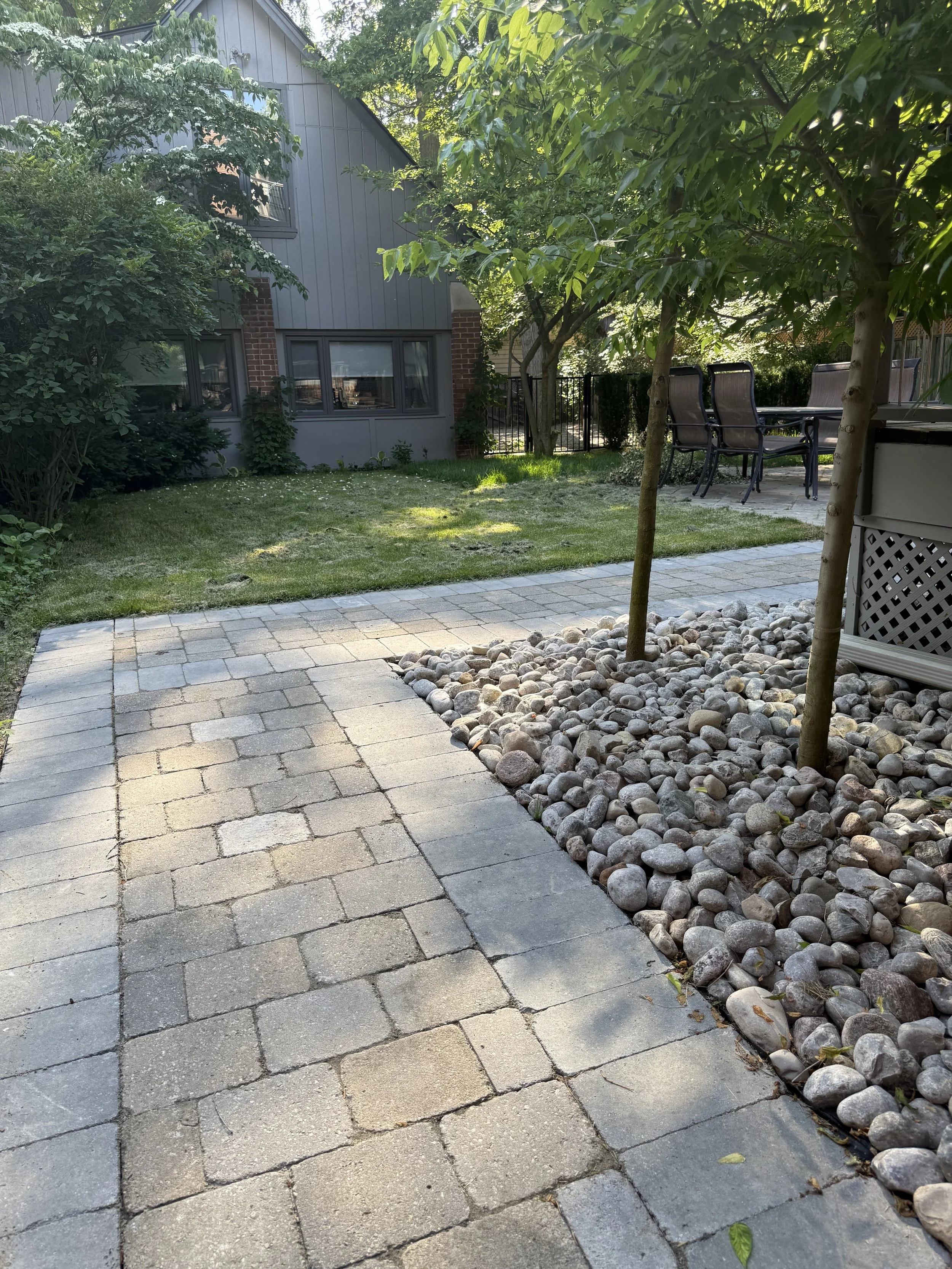 A backyard with a stone patio, edged with smooth pebbles, trees, and a small lawn. There is outdoor furniture, including a table and chairs, near a fence. A gray house with multiple windows is visible in the background.