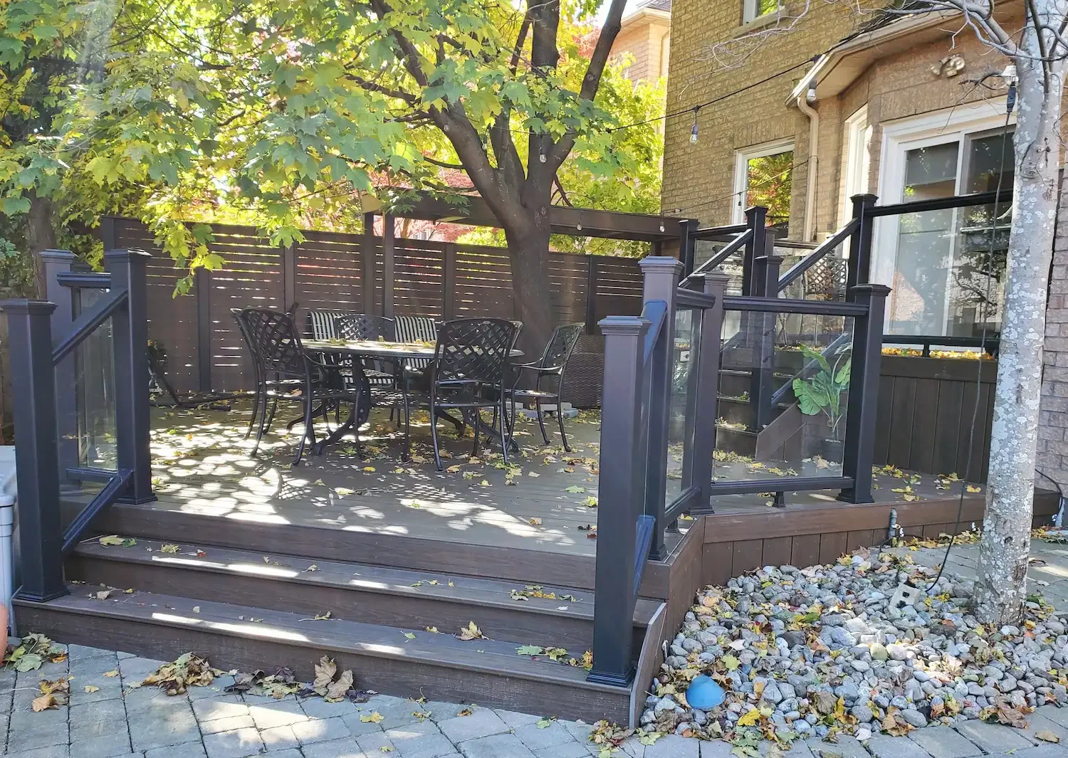 Backyard patio with black metal outdoor furniture, a tree with green leaves, and a charcoal-colored deck with a railing. There are stairs leading down from the deck, and a small garden area with rocks and a tree trunk on the right side.