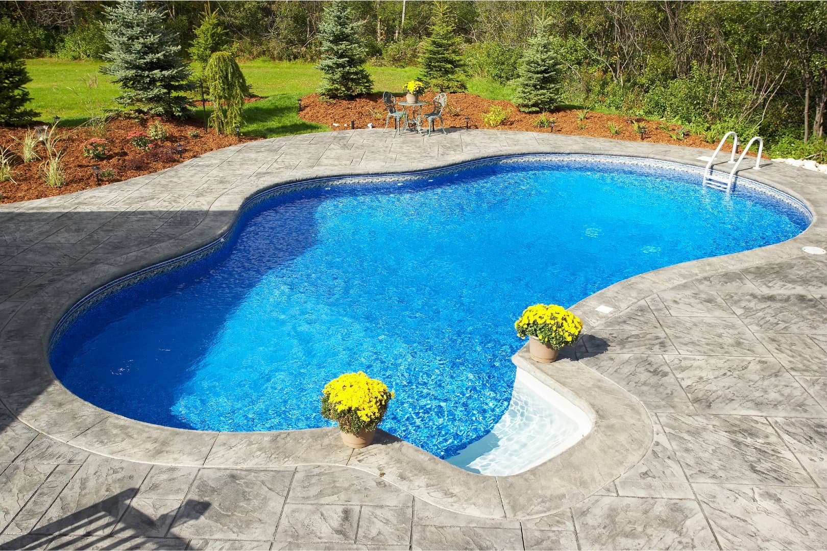 In-ground swimming pool with a curved shape, surrounded by a stone patio, with two yellow potted flowers at the pool's edge. In the background, there is greenery with trees and shrubs, and a small outdoor seating area with a table and chairs.