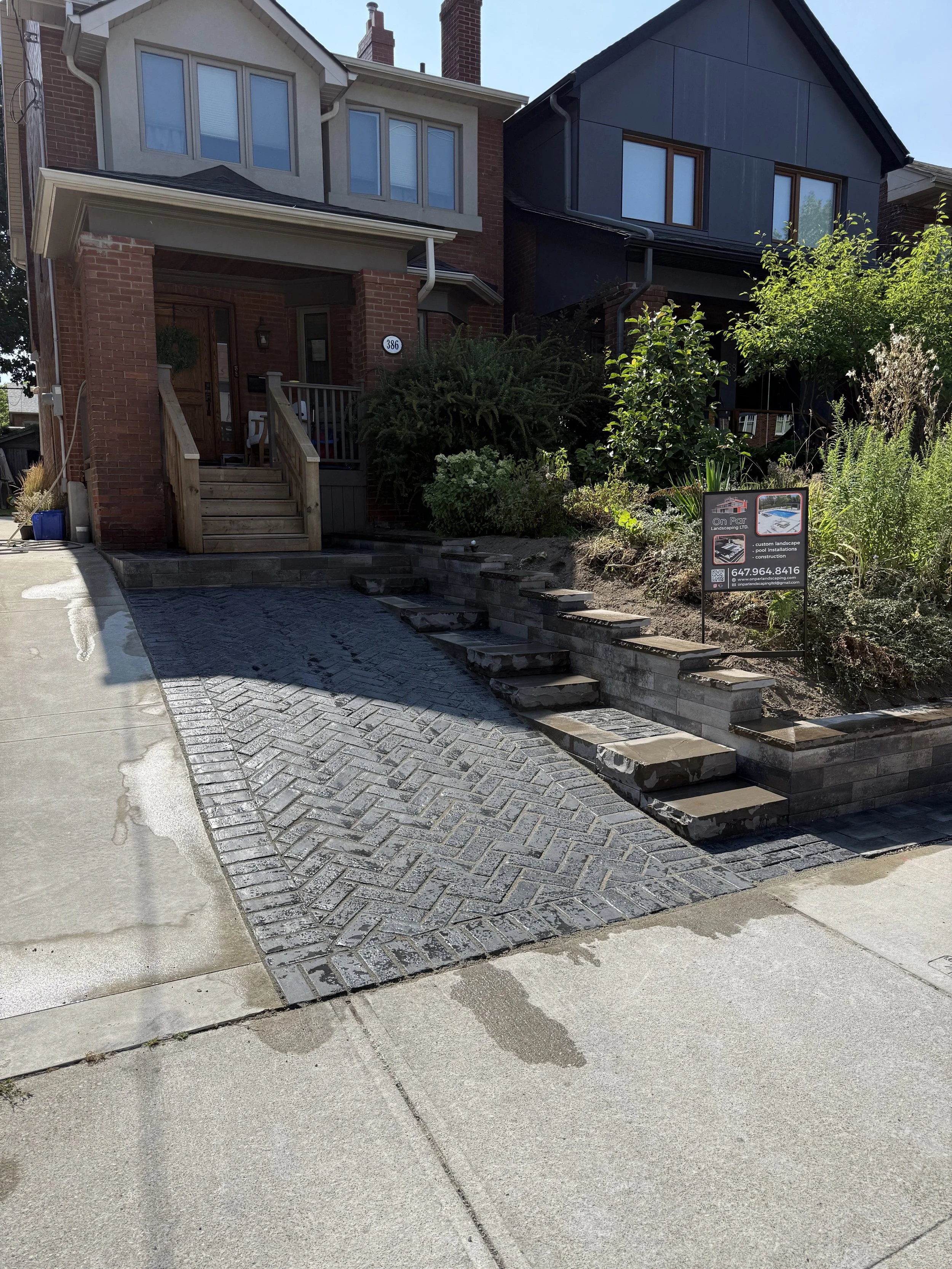 A residential house with a driveway paved with gray bricks and a stone staircase leading to the front porch. There is a small sign on the garden bed with landscaping company information.