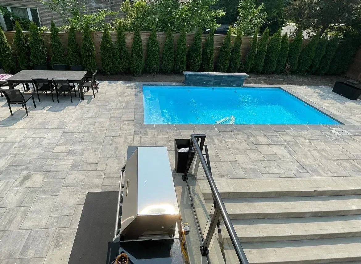 Backyard with outdoor dining table and chairs, swimming pool with water, and a paved patio surrounded by green shrubs and trees.