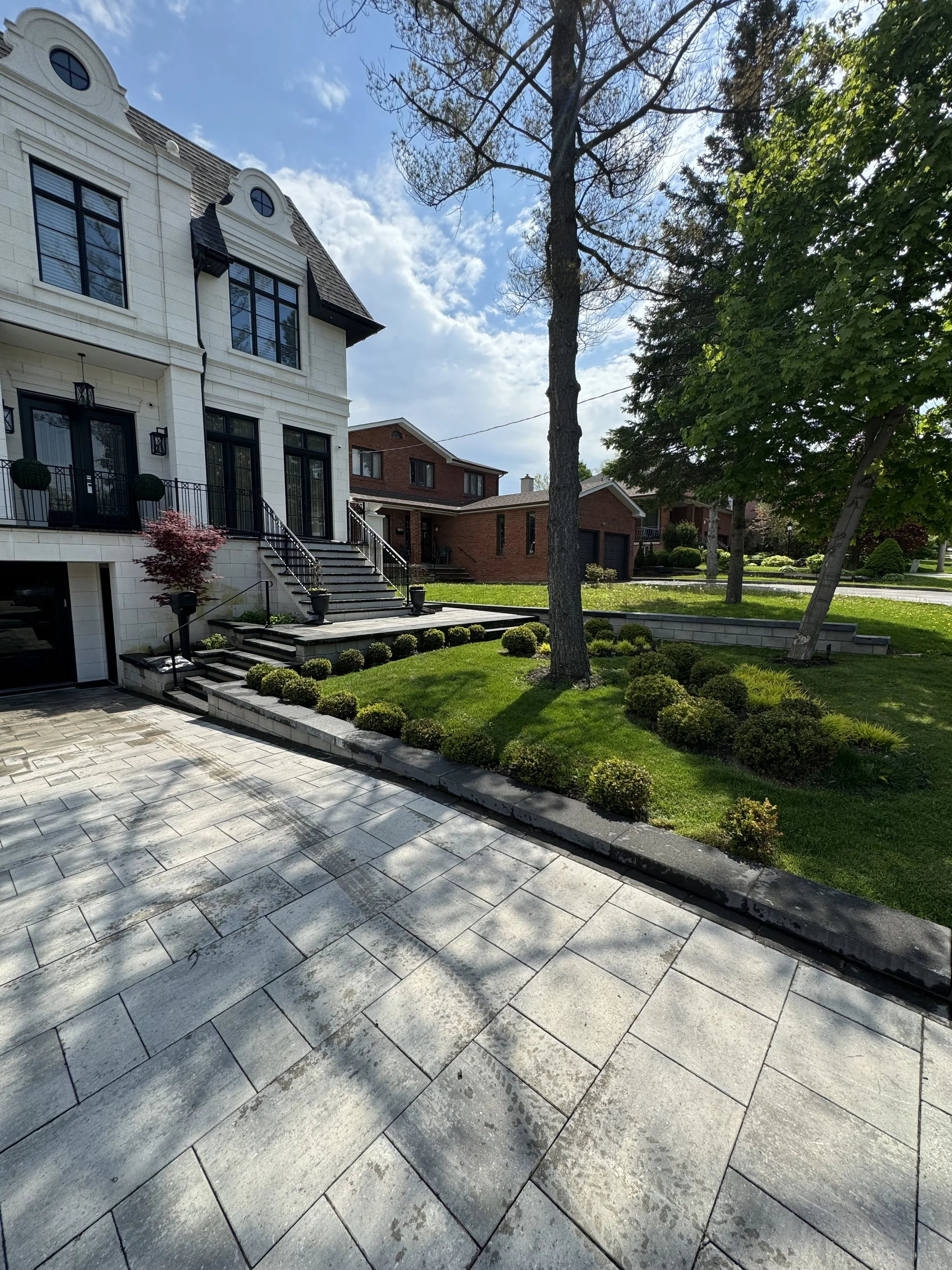A modern two-story house, a well-maintained lawn, and landscaped shrubs. There is a paved driveway and steps leading to the entrance. A tree and clear blue sky are visible in the background.