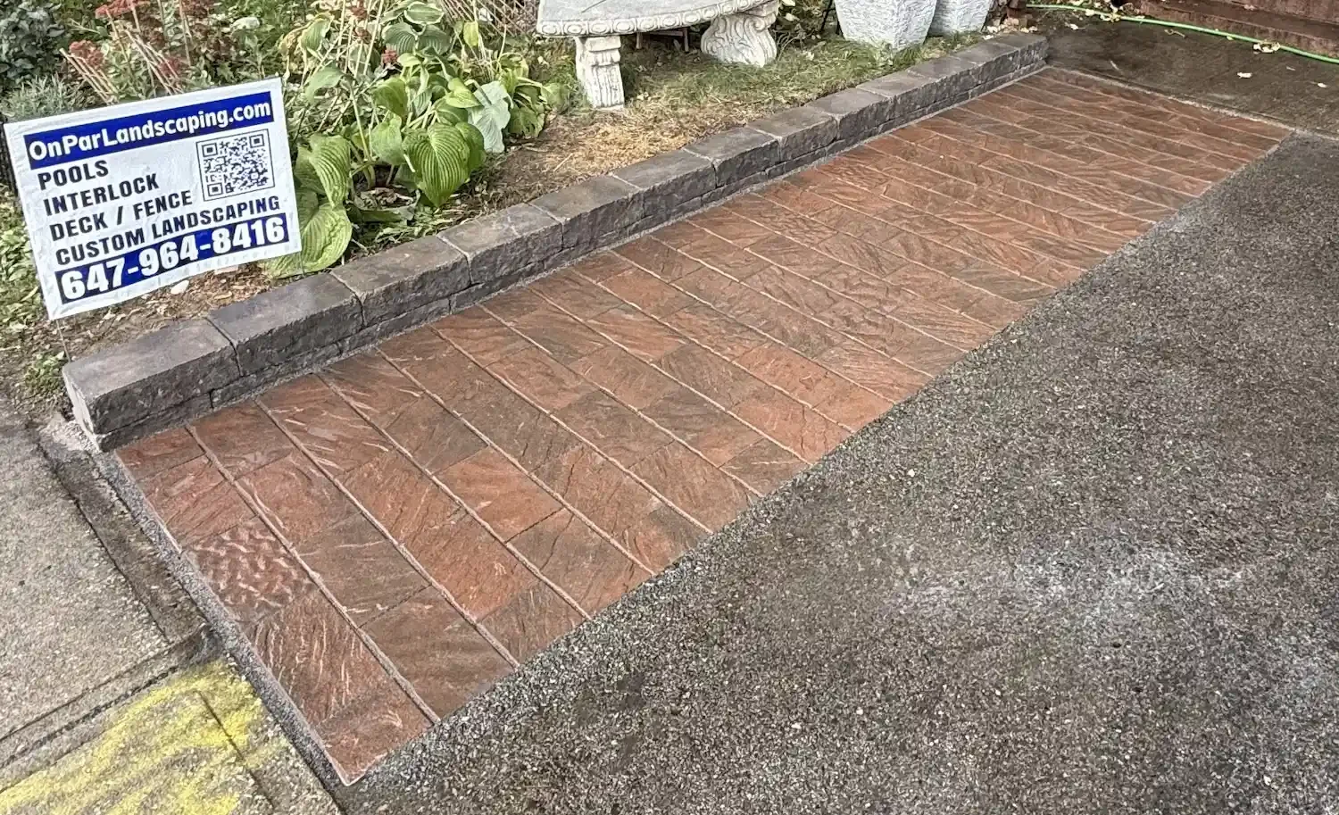 A newly paved brick pathway with a dark gray stone border on a lawn area, next to a garden with green plants, and a sign advertising landscaping services.