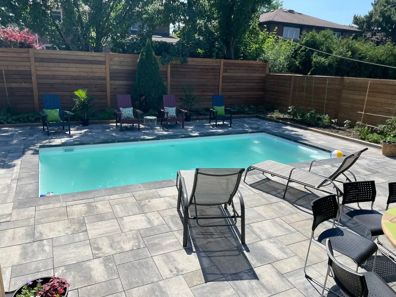 A backyard with a swimming pool, outdoor chairs, and potted plants, enclosed by a wooden fence.