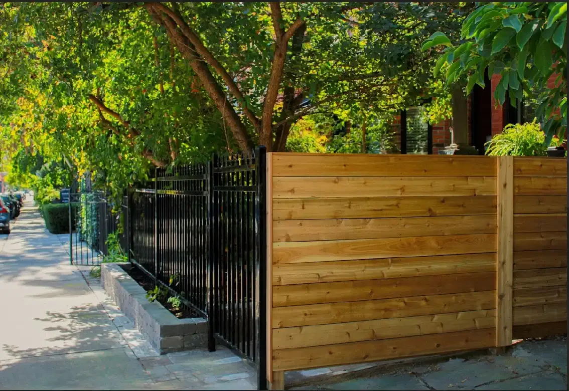 View of a sidewalk with a black iron fence and wooden privacy fence beside a tree, with parked cars and greenery in the background.