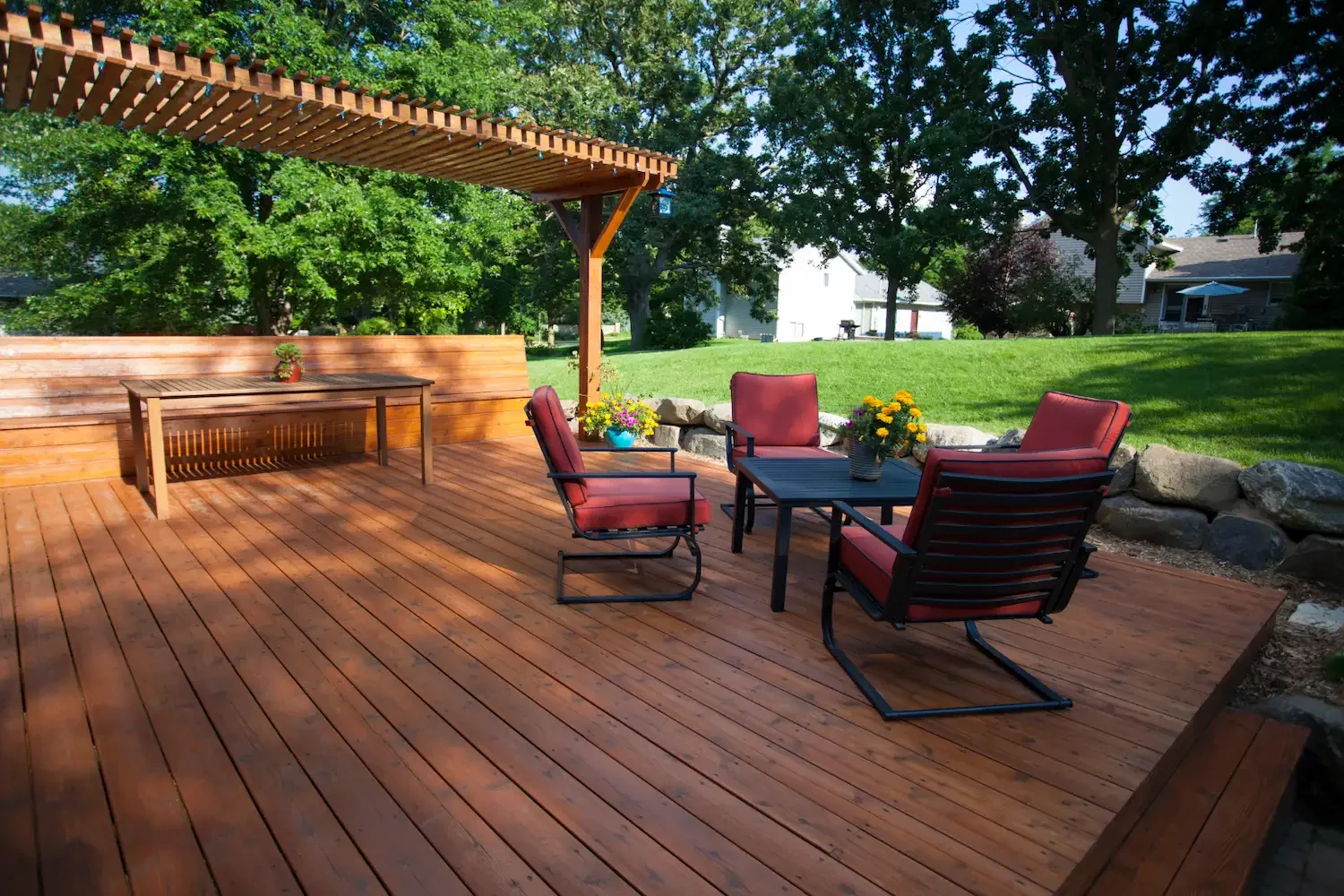 A backyard wooden deck with outdoor furniture, including four red cushioned chairs around a small black table with yellow flowers, a wooden bench, and a wooden table with a small potted plant, shaded by a large tree and a wooden pergola, surrounded b