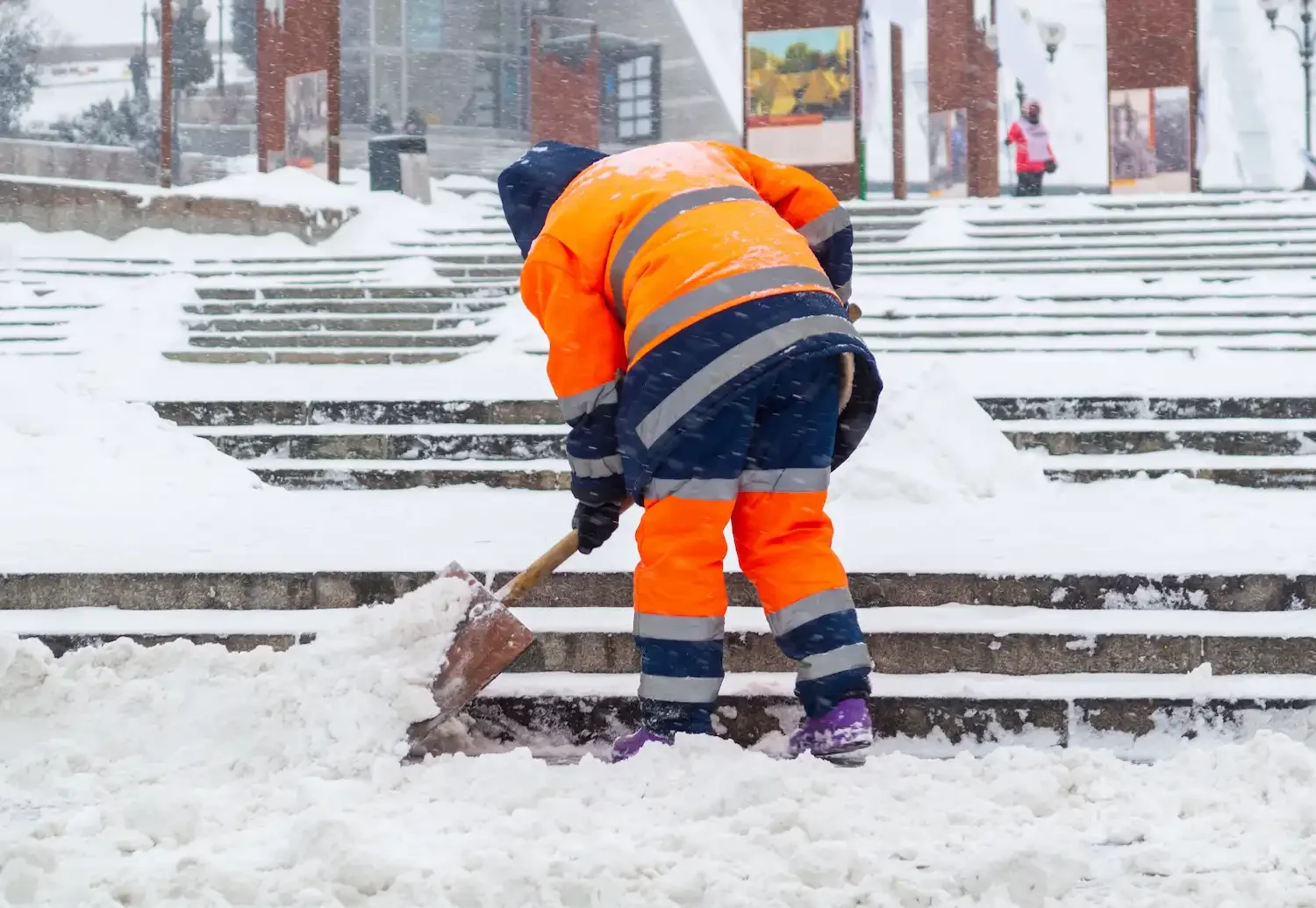 A person dressed in a bright orange and navy reflective winter coat and pants shoveling snow on concrete stairs during a snowstorm. The person is wearing purple shoes, and there are buildings and another person in the background.