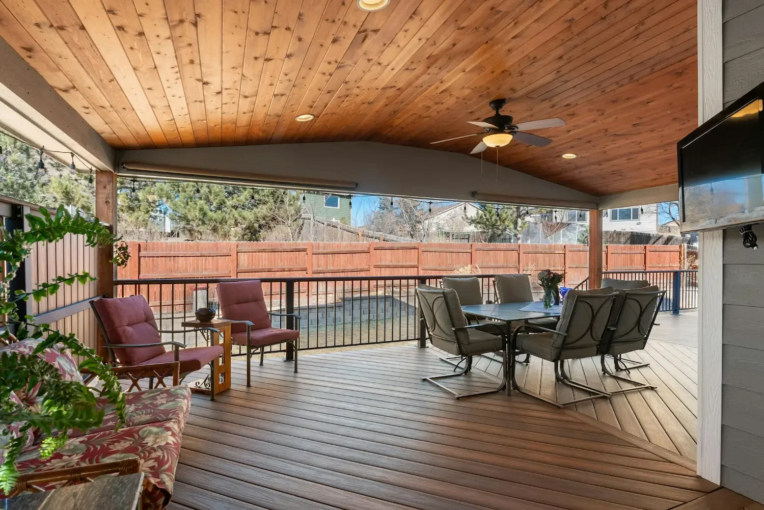 Covered backyard patio with wooden ceiling, ceiling fan, and outdoor furniture including a dining table with six chairs, and several cushioned chairs along the railing. There is a patio TV mounted on the wall and a string of outdoor lights hanging ab
