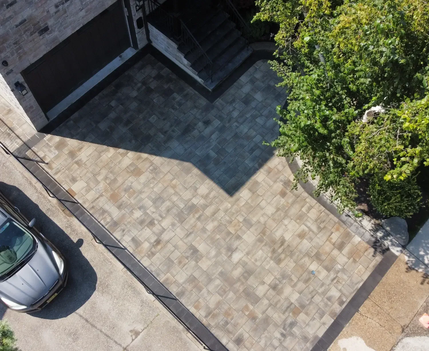 An aerial view of a concrete paved courtyard next to a building and a parked silver car. There are trees on one side providing shade.