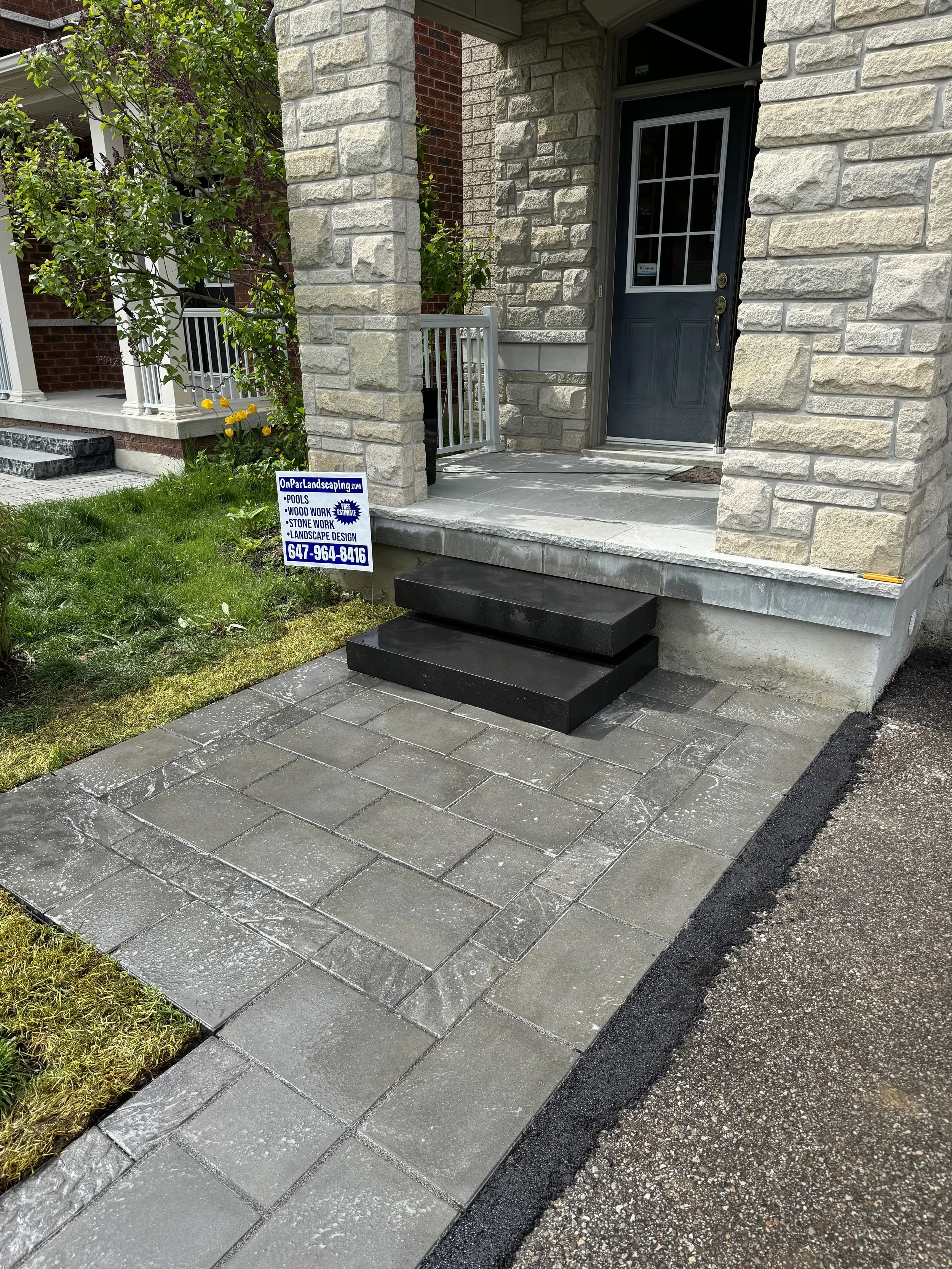 Front porch of a house with stone steps and a black door. Newly laid grey stone pavement leading to the steps, with a small lawn and a sign for landscaping services.