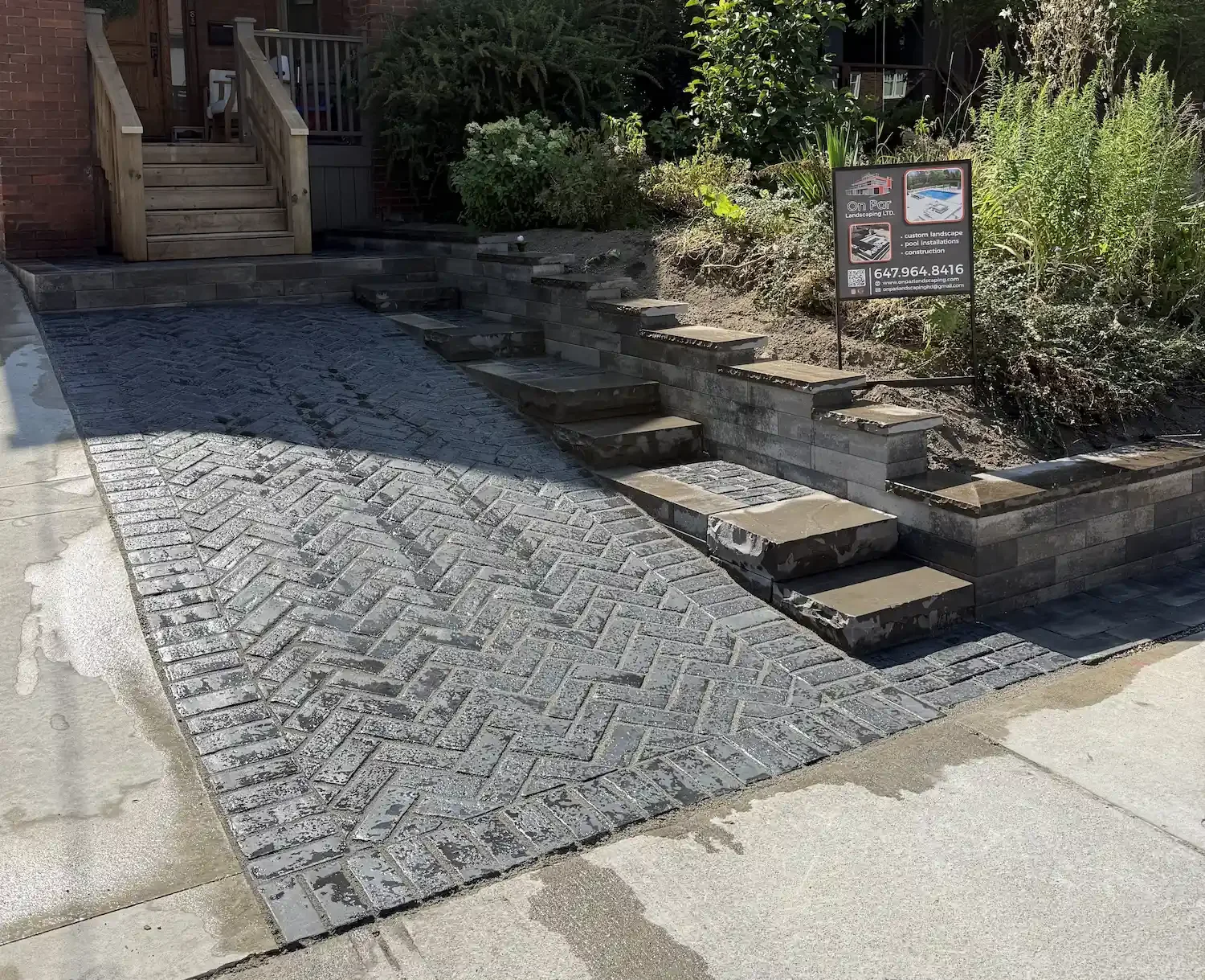 A newly constructed outdoor landscape featuring a herringbone brick driveway, a set of stone steps, and a layered stone retaining wall with a garden area and a sign promoting custom landscape and pool installations.