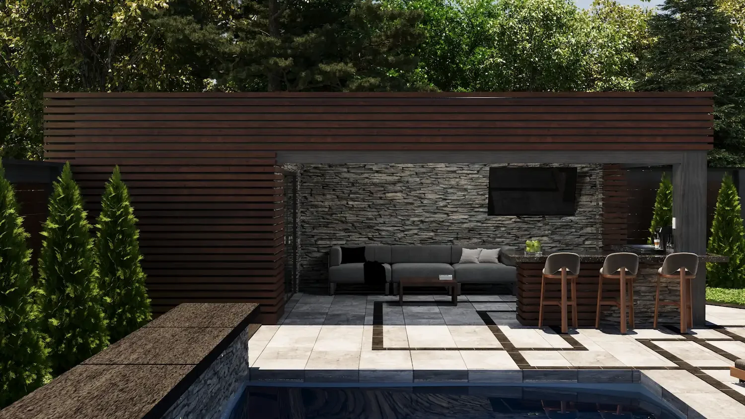 Outdoor patio with a stone wall, a gray sectional sofa with white pillows, a flat-screen TV on the wall, a bar area with four barstools, a pool in the foreground, and lush green trees in the background.