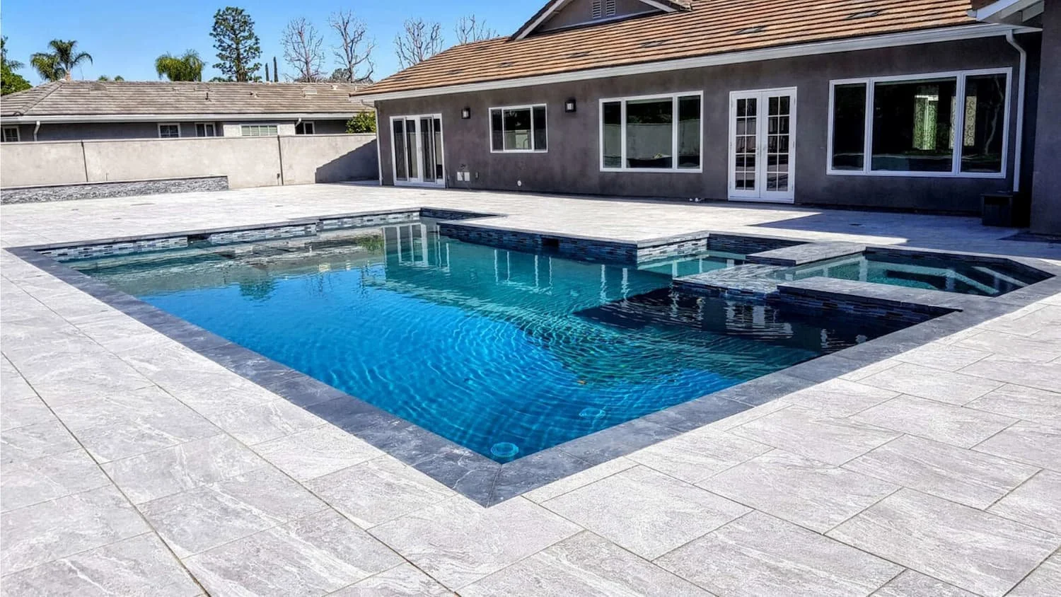 Backyard with a rectangular swimming pool featuring blue water, surrounded by a light gray stone deck, with a house in the background that has sliding glass doors and multiple windows.