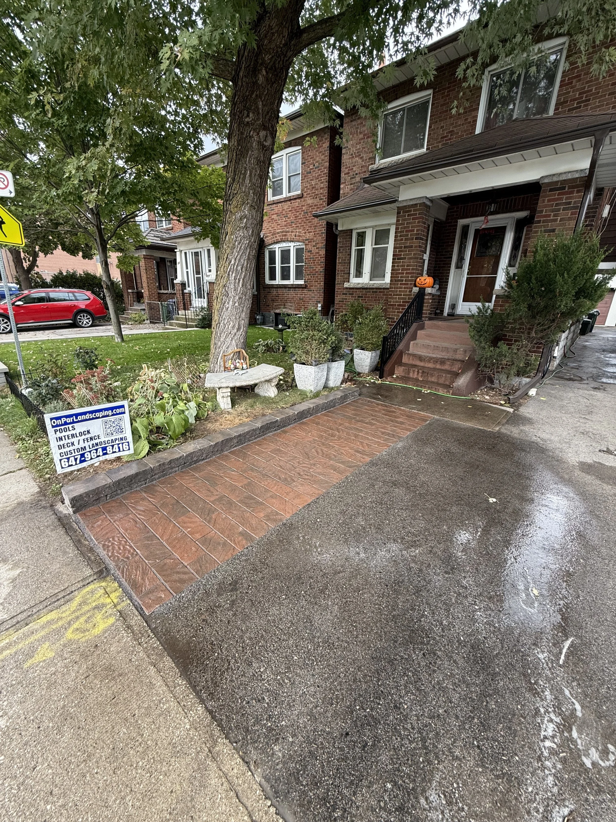 Front yard of a brick rowhouse with a small landscaped garden, a tree, a wooden bench, potted plants, Halloween pumpkin decoration, and a newly laid brick and concrete driveway.