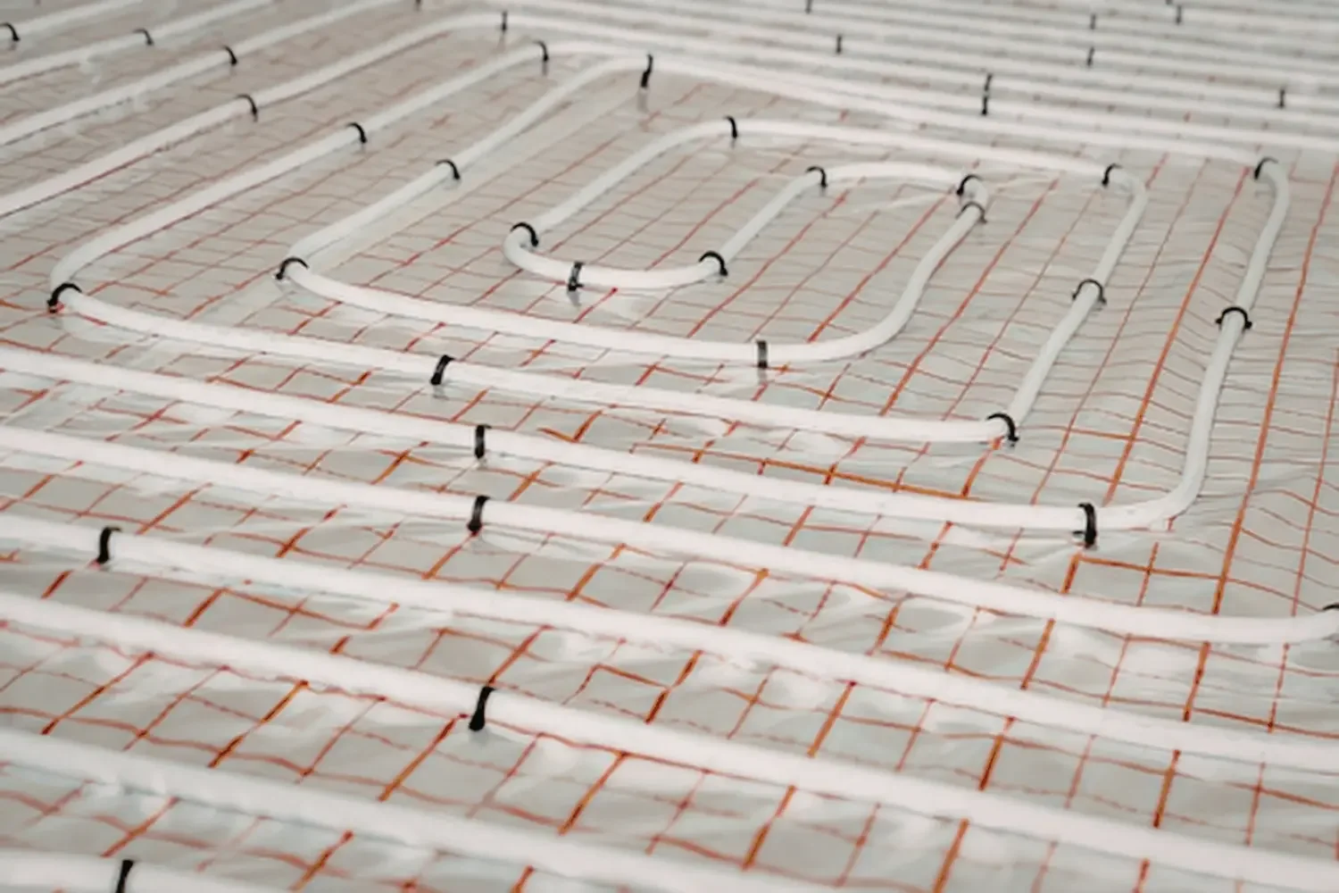 Underfloor heating system with white pipes installed in a concrete floor, held down by black clips and laid on a grid of red mesh.